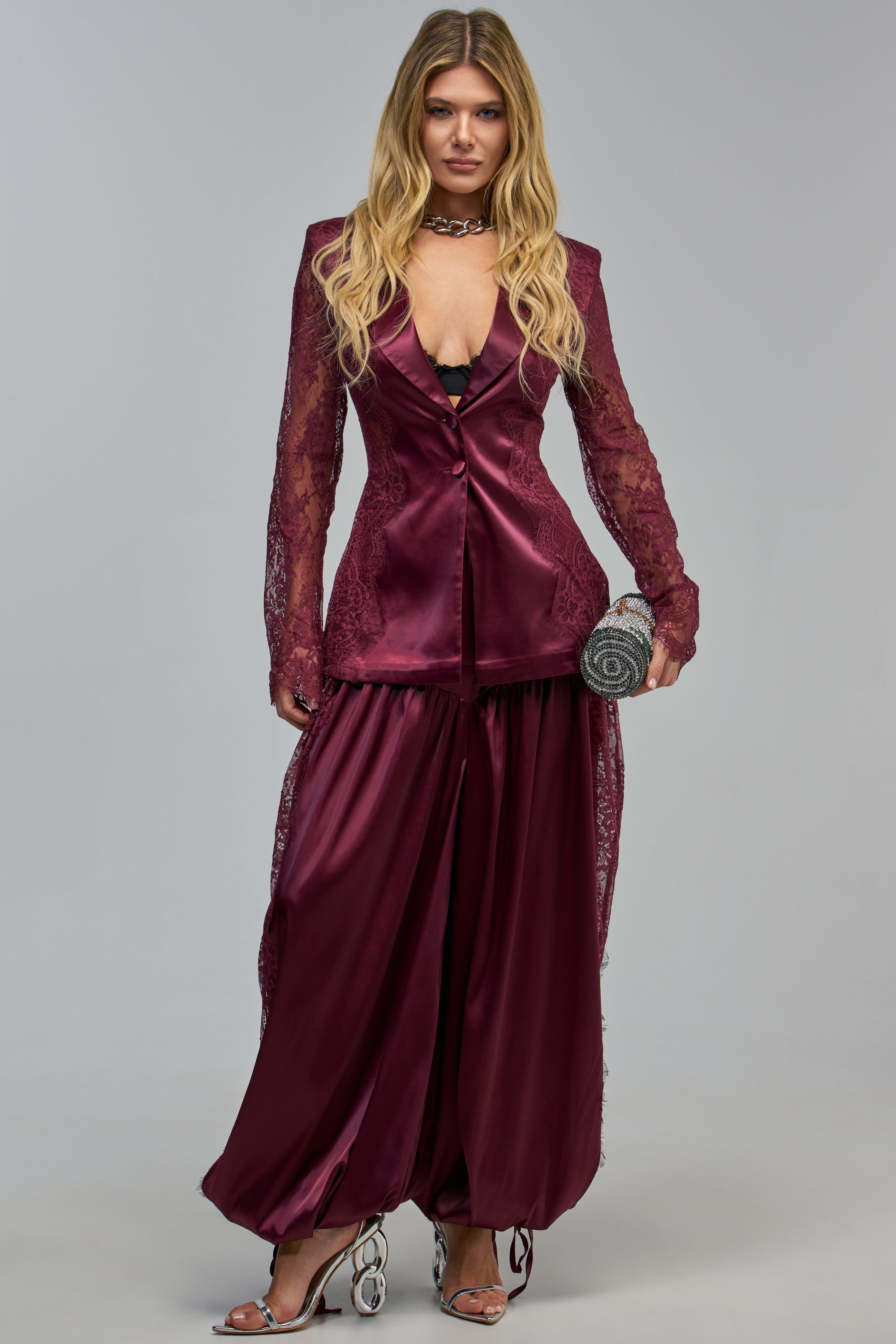 A person wearing a burgundy satin and lace blazer with matching AU CONTRAIRE LACE SATIN BALLOON PANTS IN BURGUNDY, holding a circular embellished clutch.