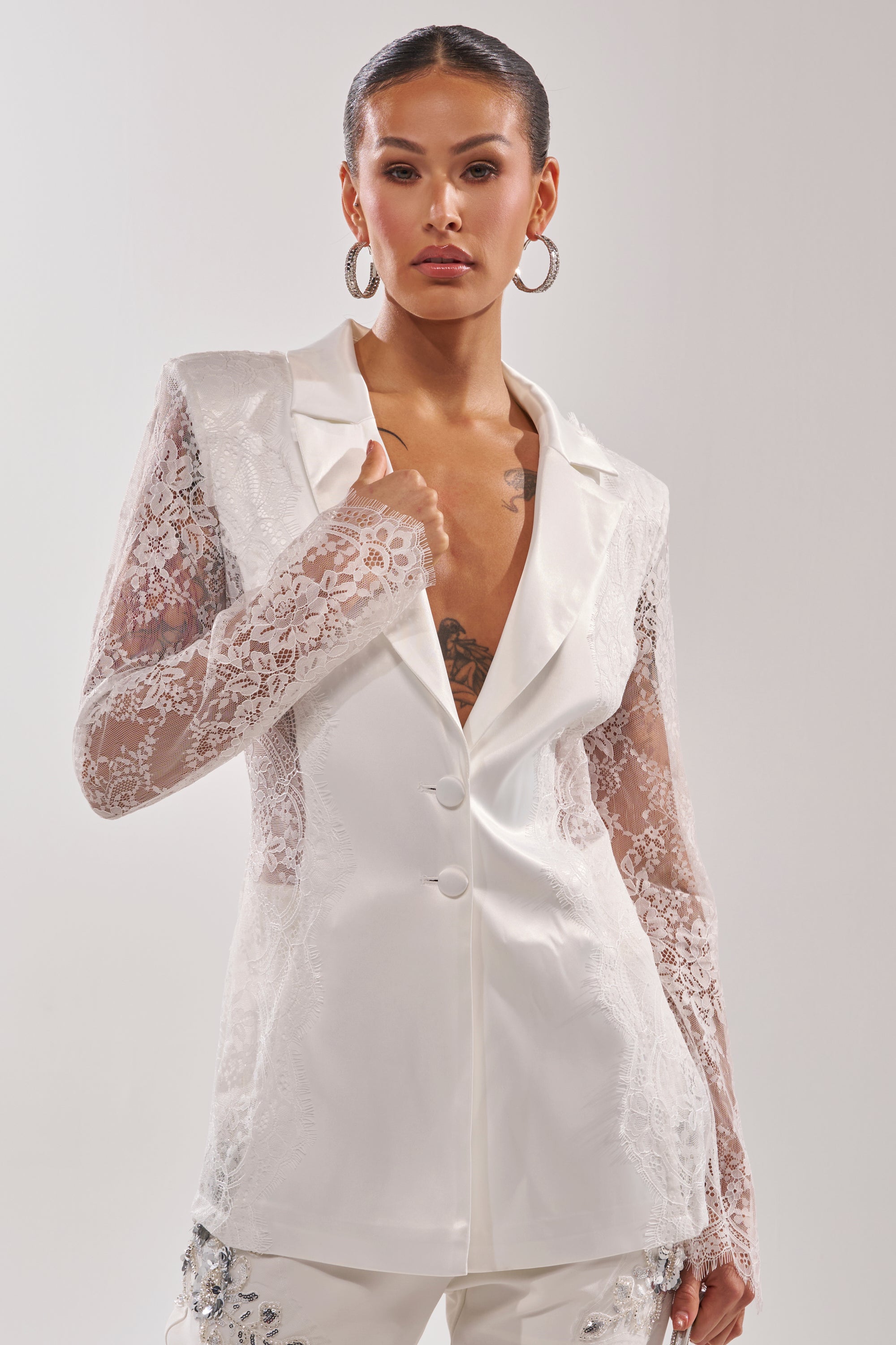 A woman stands confidently against a plain background wearing the AU CONTRAIRE LACE SATIN BLAZER IN WHITE, featuring sheer lace sleeves and lapels, paired with large hoop earrings and embellished white pants. 1