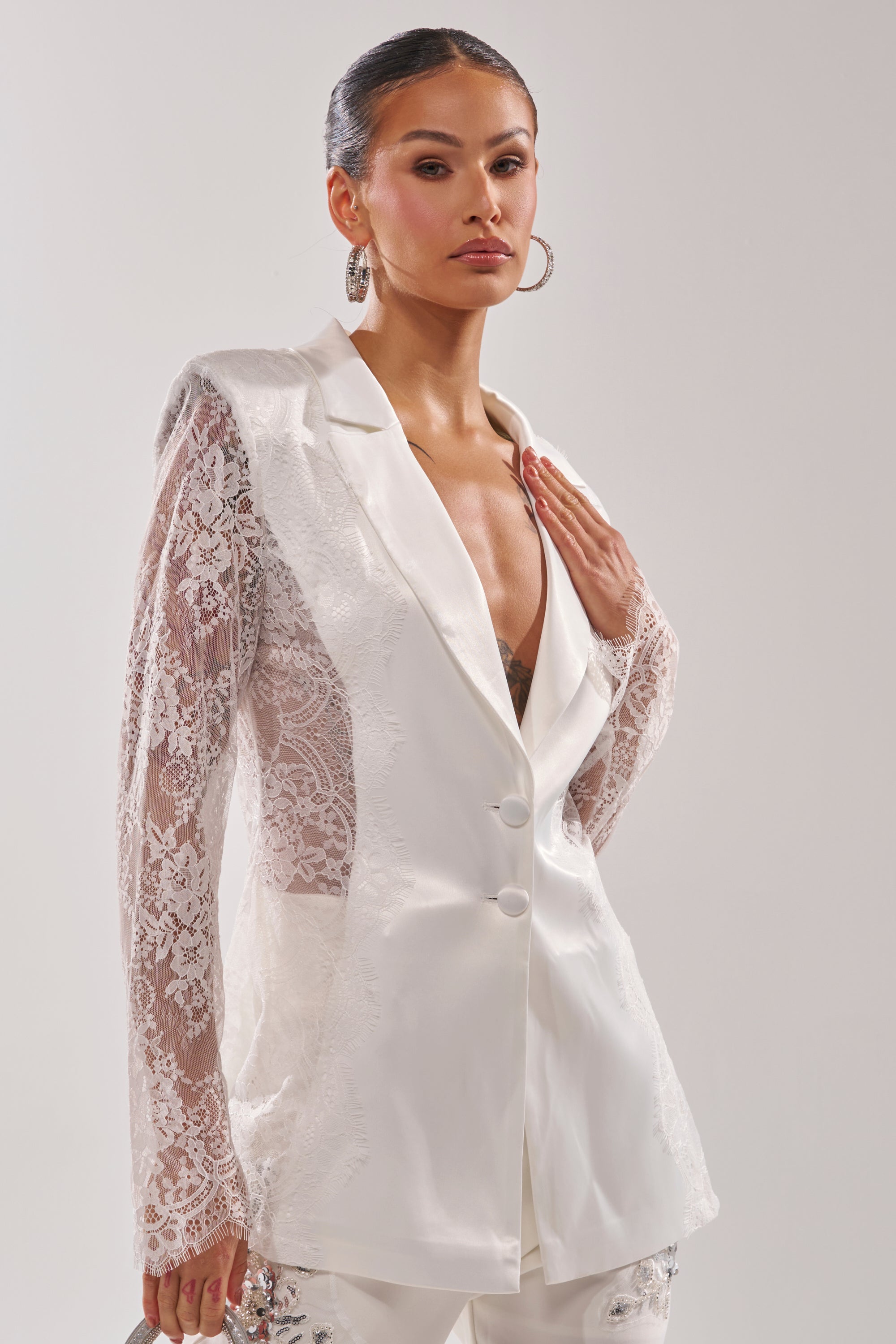 A woman poses confidently against a plain light background wearing the AU CONTRAIRE Lace Satin Blazer in White, featuring tailored details and lace sleeves, paired with large hoop earrings. 2