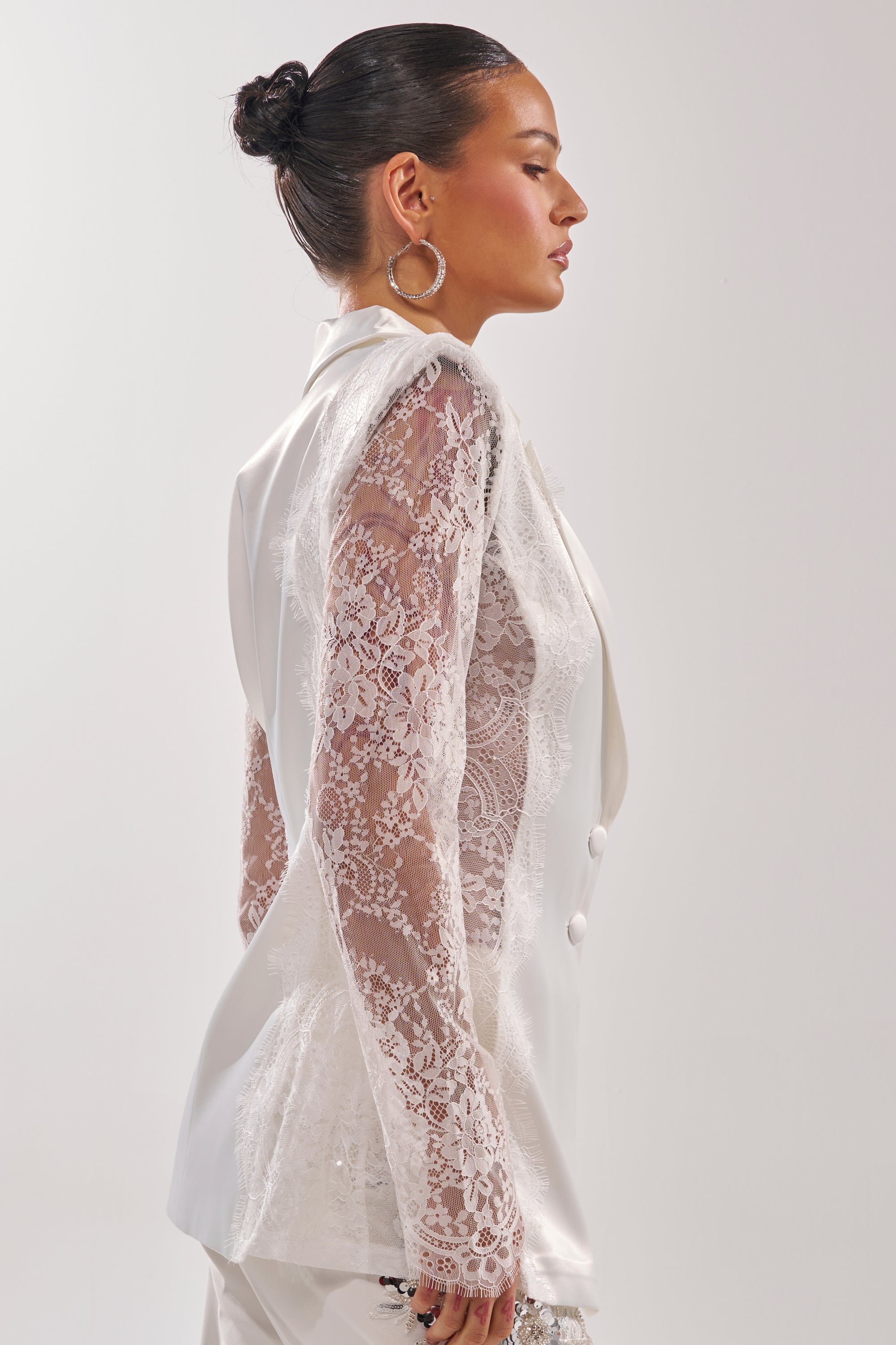 A woman with dark hair in a sleek bun wears the AU CONTRAIRE LACE SATIN BLAZER IN WHITE, featuring sheer lace sleeves and large hoop earrings, shown in profile against a plain light background. 3