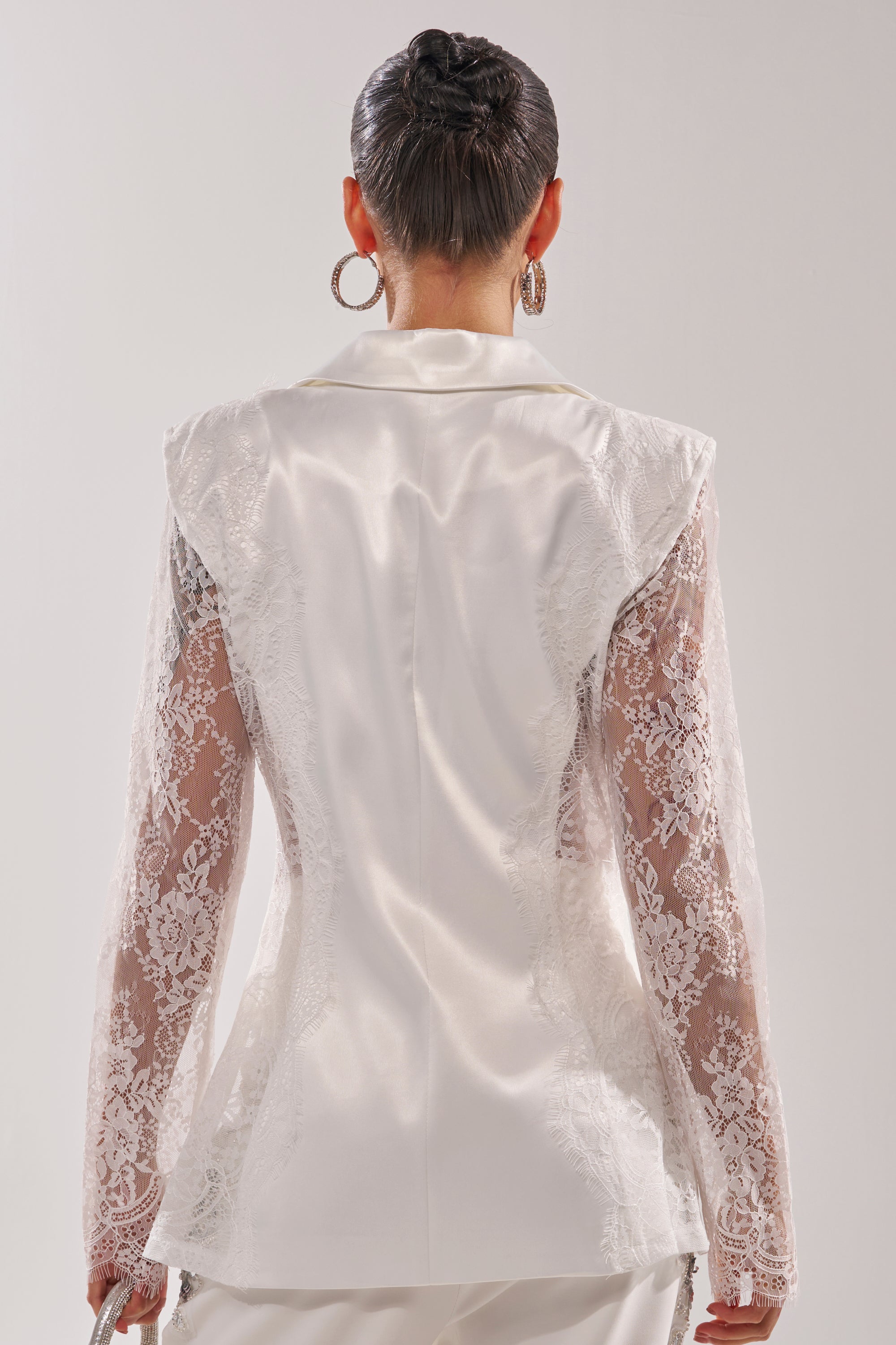 A woman faces away, wearing the AU CONTRAIRE LACE SATIN BLAZER IN WHITE with sheer lace sleeves and structured shoulders. Her hair is styled in a sleek bun, complemented by large hoop earrings. 4