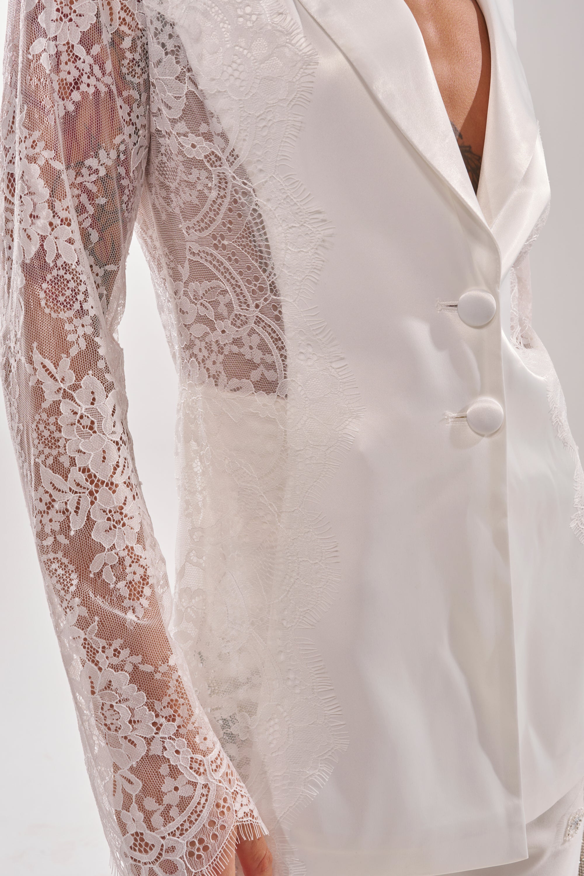 A close-up of the AU CONTRAIRE LACE SATIN BLAZER IN WHITE: a white tailored blazer with sheer floral lace sleeves and lapels, two white buttons, and a deep V-neck—blending intricate elegance with modern style. 7