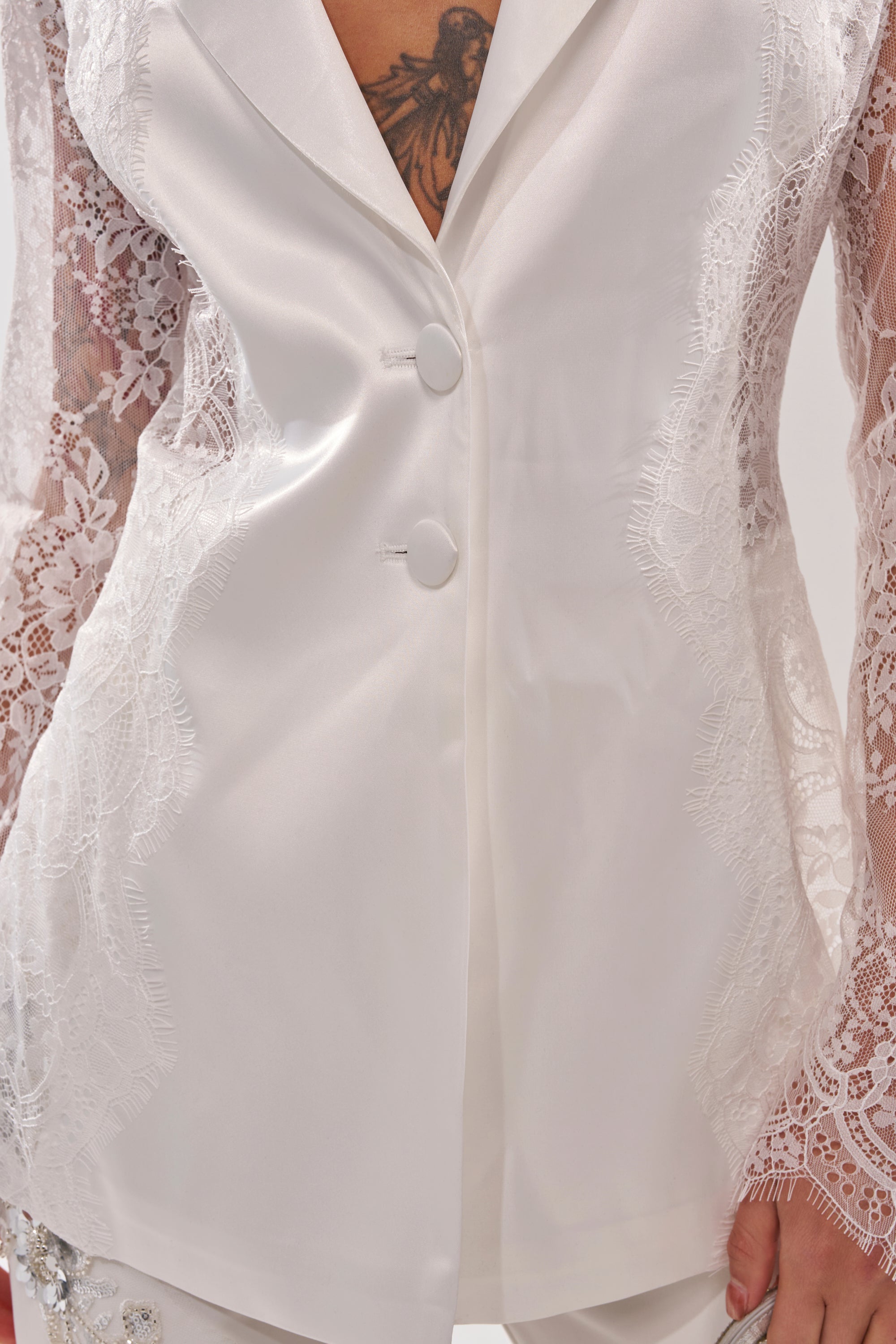 A close-up of a person wearing the AU CONTRAIRE LACE SATIN BLAZER IN WHITE, featuring lace sleeves, intricate detailing, and two front buttons. A chest tattoo is partially visible above the blazer's neckline. 8