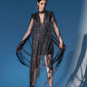 A woman poses confidently against a bold blue backdrop, wearing the MIXED MESSAGES METALLIC BANDAGE MINI DRESS WITH FRINGE. The shimmering black mini dress features dramatic fringe from her shoulders and arms, paired with high heels and statement earrings.