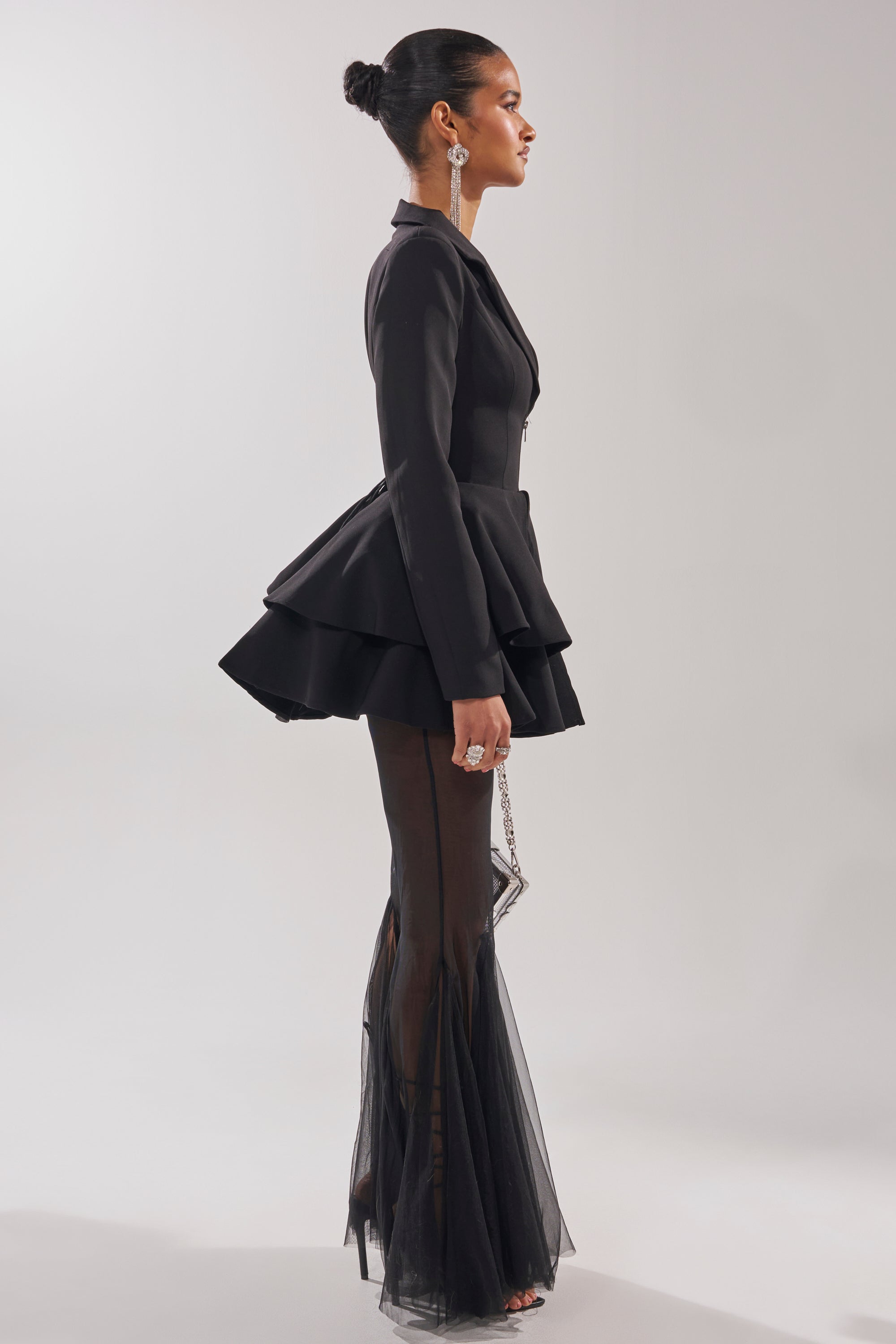 A woman stands in profile against a plain background, wearing the WORK HARD PLAY HARD BLAZER MESH JUMPSUIT featuring a peplum, short skirt overlay, and sheer flared pants—her bun hairstyle and statement earrings complete this striking formal look.
