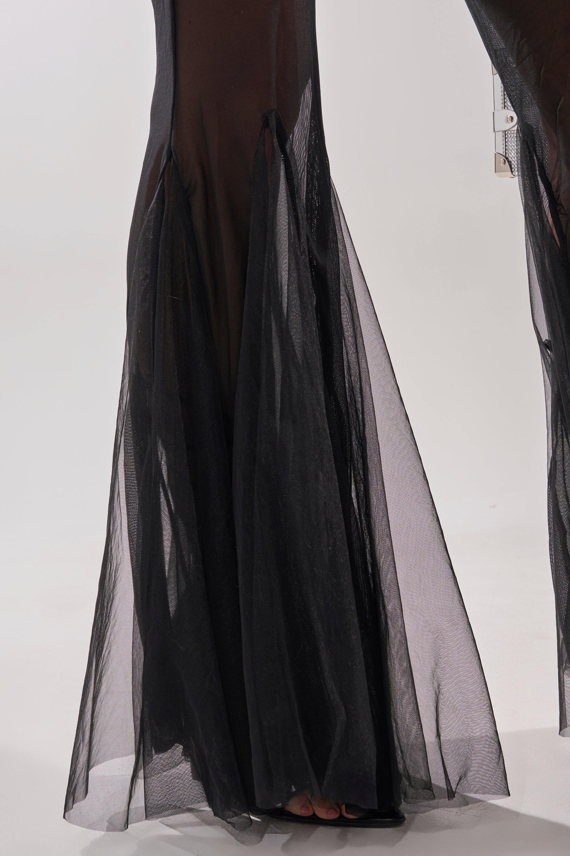 A close-up of the lower half of the WORK HARD PLAY HARD BLAZER MESH JUMPSUIT, featuring black sheer mesh with flowing bell-bottom legs and visible feet, set against a plain light background.