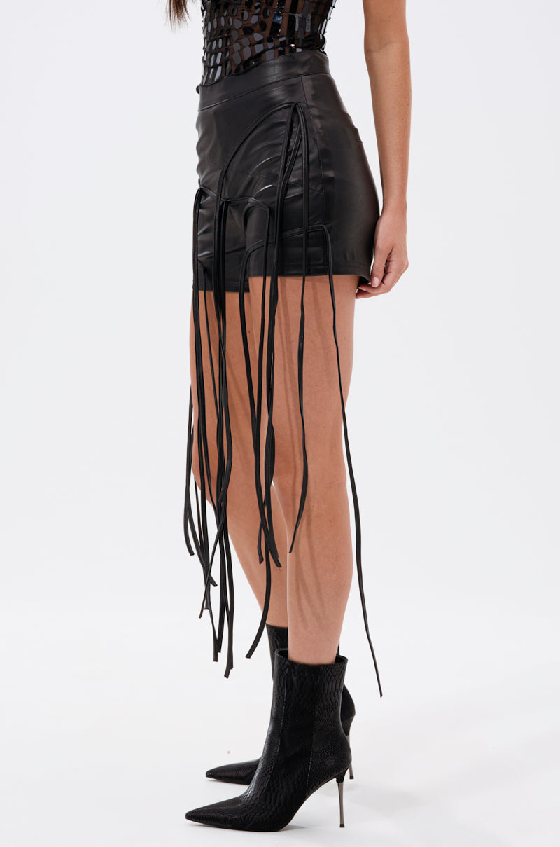 A woman wearing HANGING BY A THREAD FAUX LEATHER SHORTS with long fringe and pointed black high-heeled boots, shown from the side against a plain white background. 3