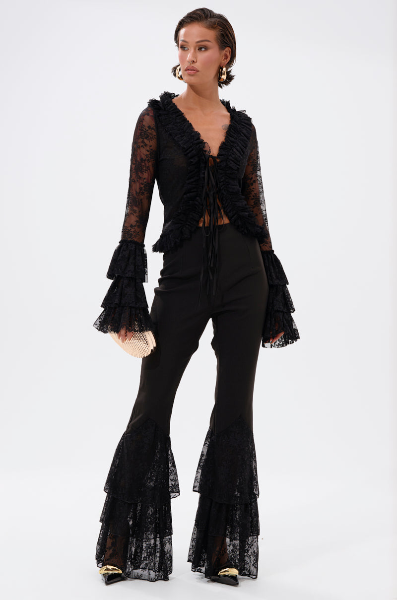 A woman models the LOOKS AND FEELS GOOD LACE FLARE LEGGING with ruffled bell sleeves, pairing it with a gold clutch, gold pointed-toe heels, slicked-back hair, and bold statement earrings for a chic look. 0