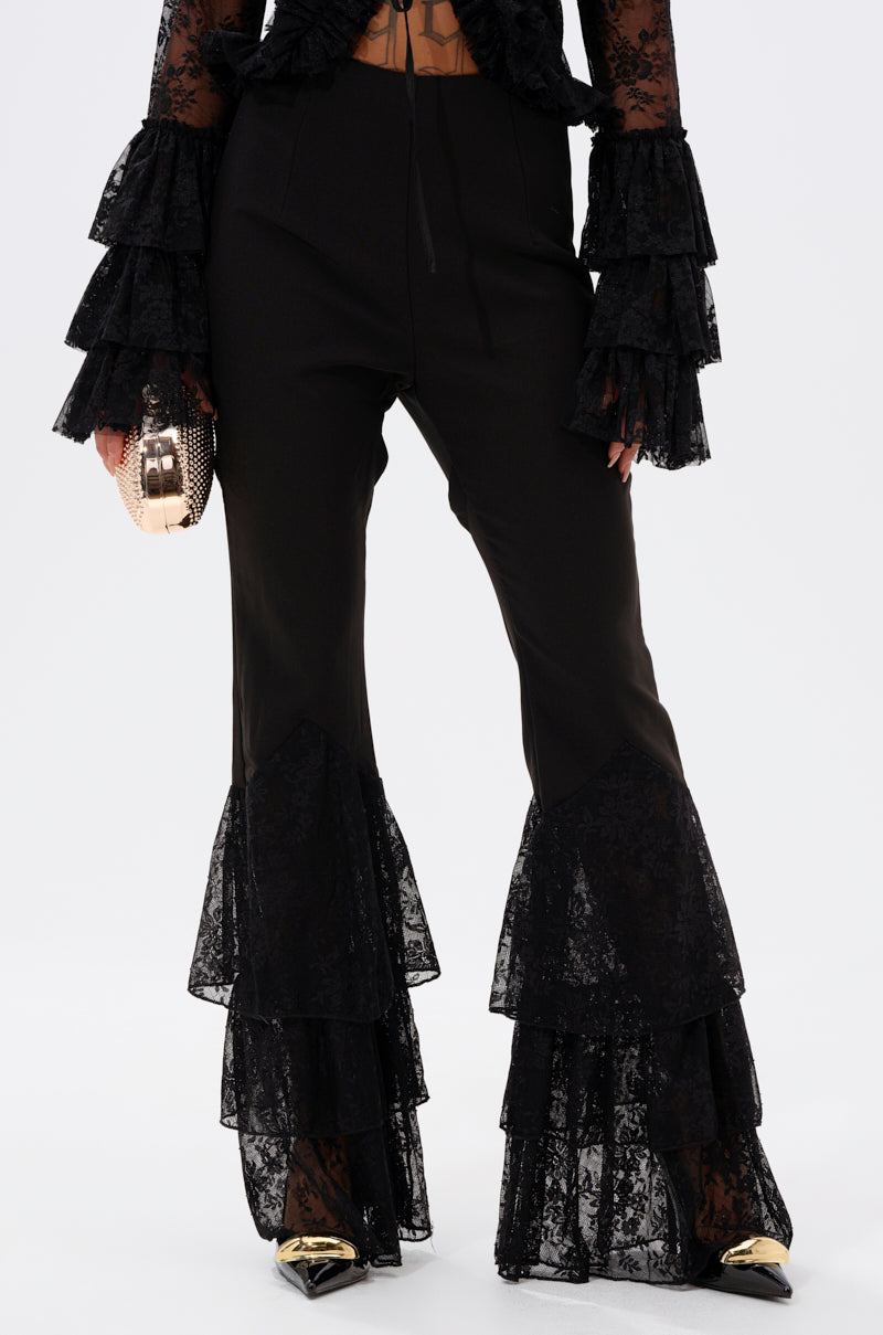 A person wearing LOOKS AND FEELS GOOD LACE FLARE LEGGING with tiered lace ruffles at the cuffs, a matching high-rise lace top with ruffled sleeves, gold pointed-toe shoes, and holding a small metallic clutch. 1