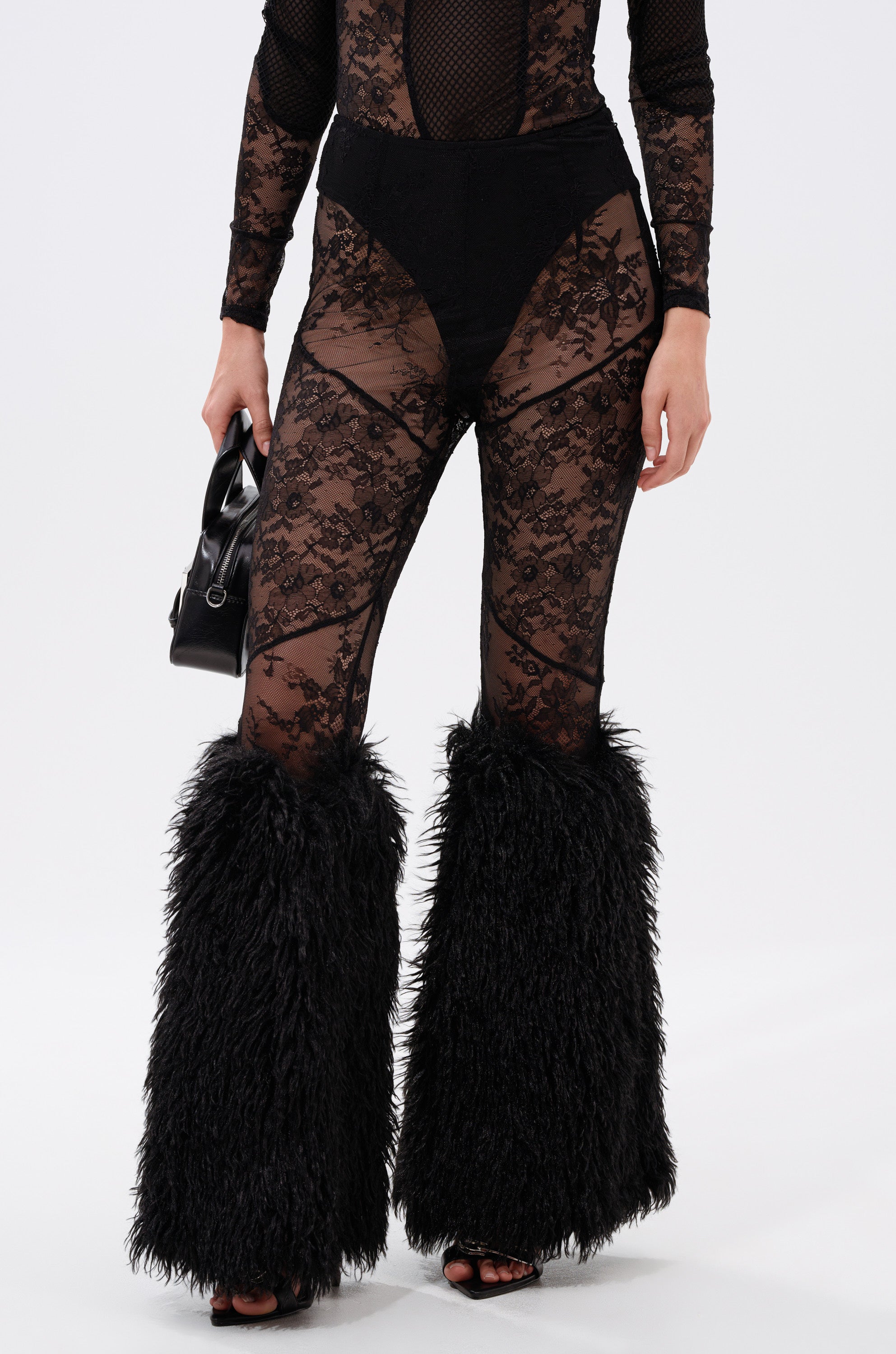 Cropped to the lower torso and legs, a person wears FEELING GOOD LACE LEGGING WITH FUR TRIM featuring a high-rise fit, sheer lace panels, prominent black faux fur trim leg warmers, and holds a small black handbag. 2
