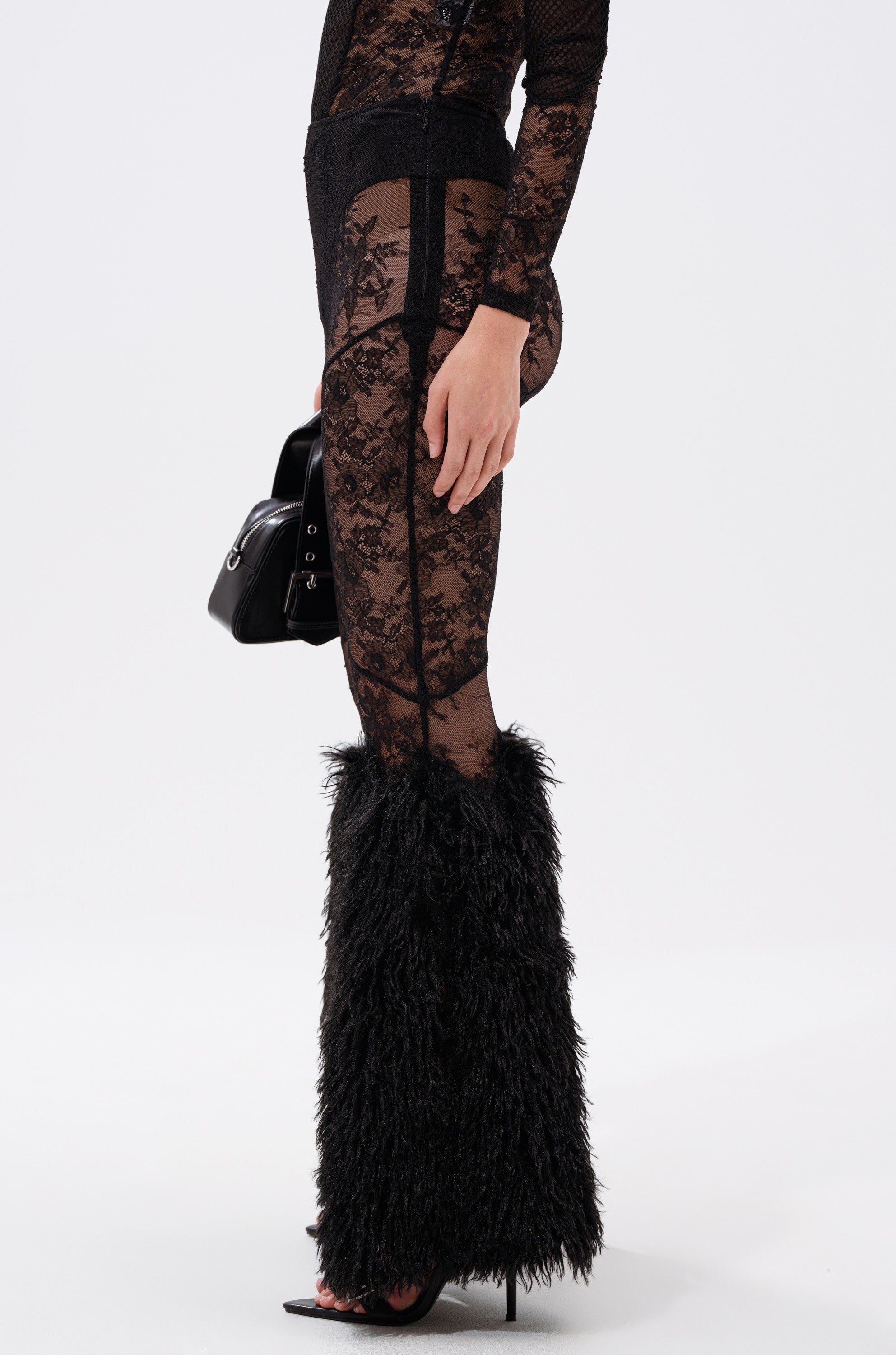 A person stands against a plain white background wearing the FEELING GOOD LACE LEGGING WITH FUR TRIM, paired with a sheer black top, furry black leg warmers, high-heeled sandals, and holding a black handbag. 4