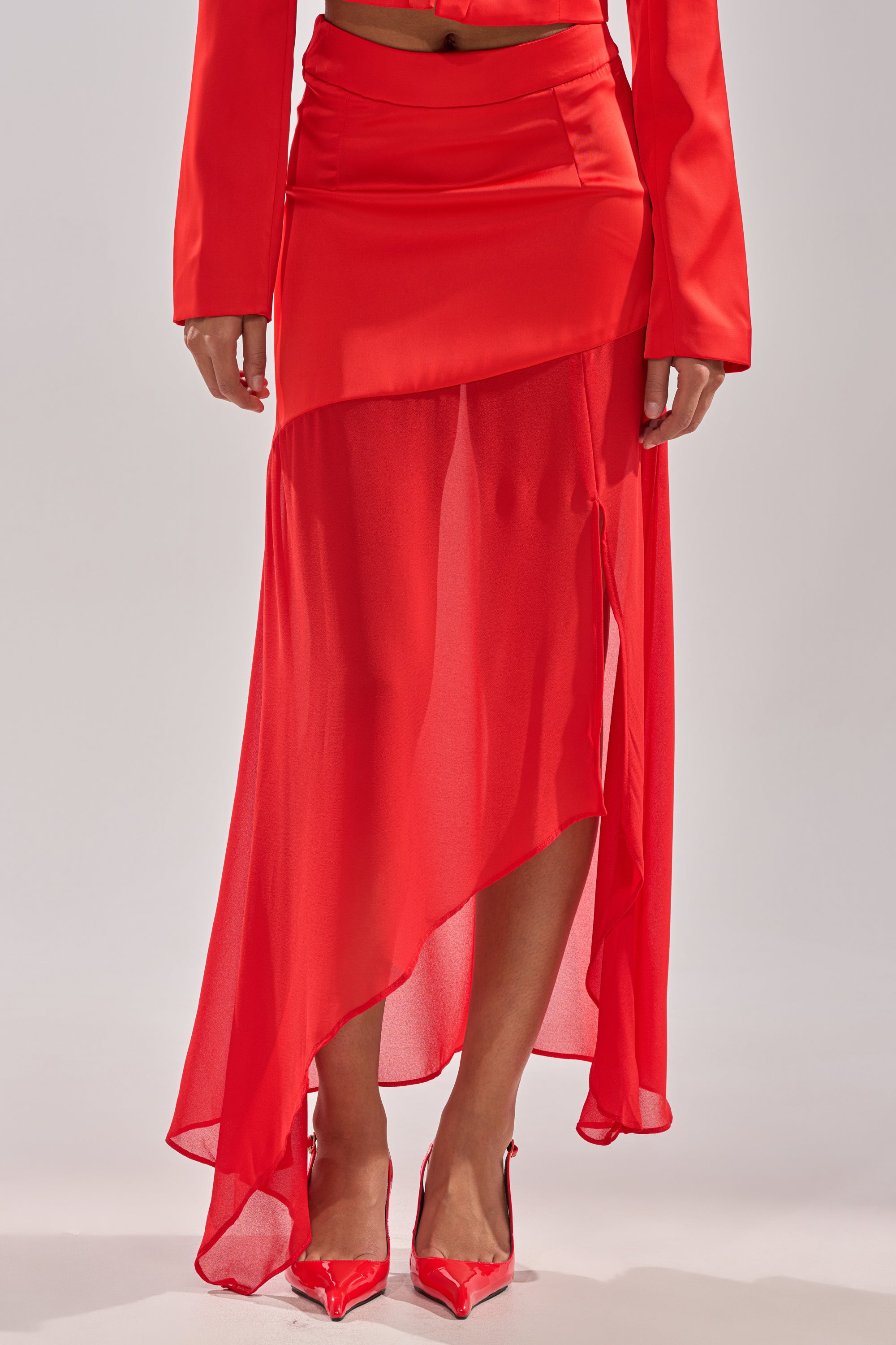 Wearing the RED HOT ASYMMETRICAL MIDI SKIRT, which features a sheer overlay atop a satin mini skirt, paired with matching red pointed heels. Only the lower half is visible for a modern, stylish look. 1
