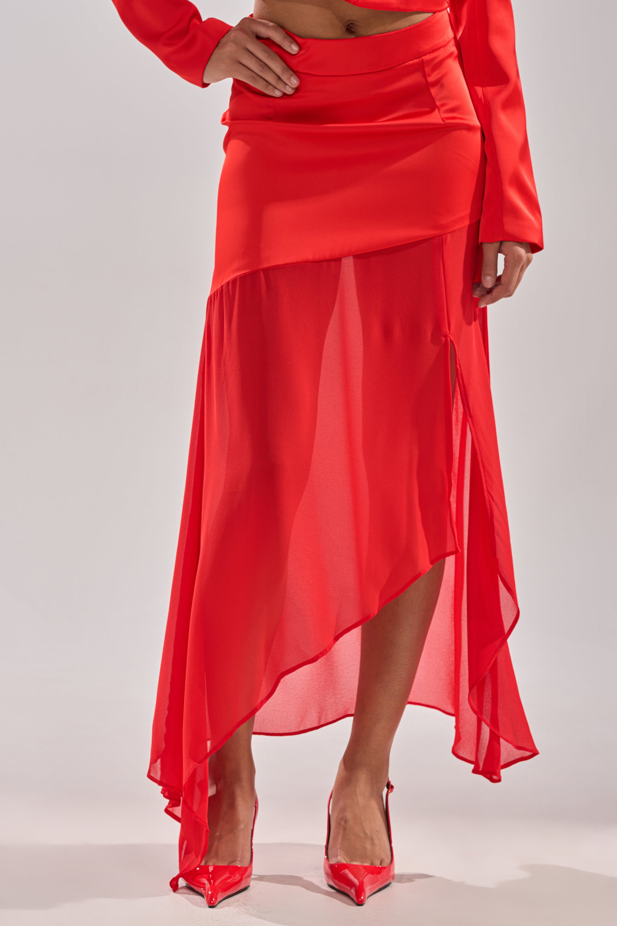 A woman poses confidently against a light background, wearing the RED HOT ASYMMETRICAL MIDI SKIRT with a high waist, paired with matching top and shiny red pointed-toe heels. 2