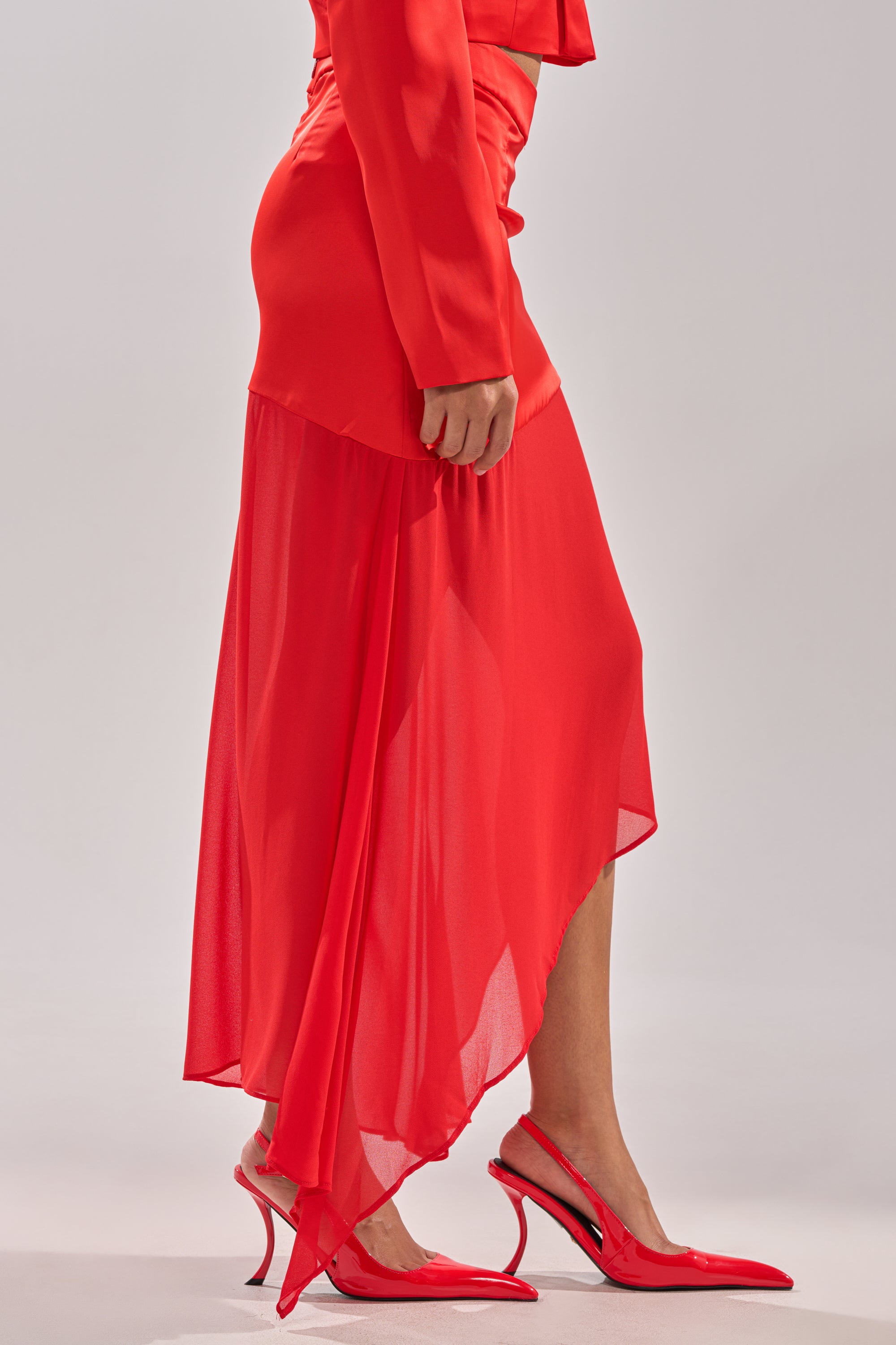 Wearing the RED HOT ASYMMETRICAL MIDI SKIRT with matching pointed-toe heels, a person stands against a plain light background, highlighting the skirt’s striking high-waist silhouette. 3