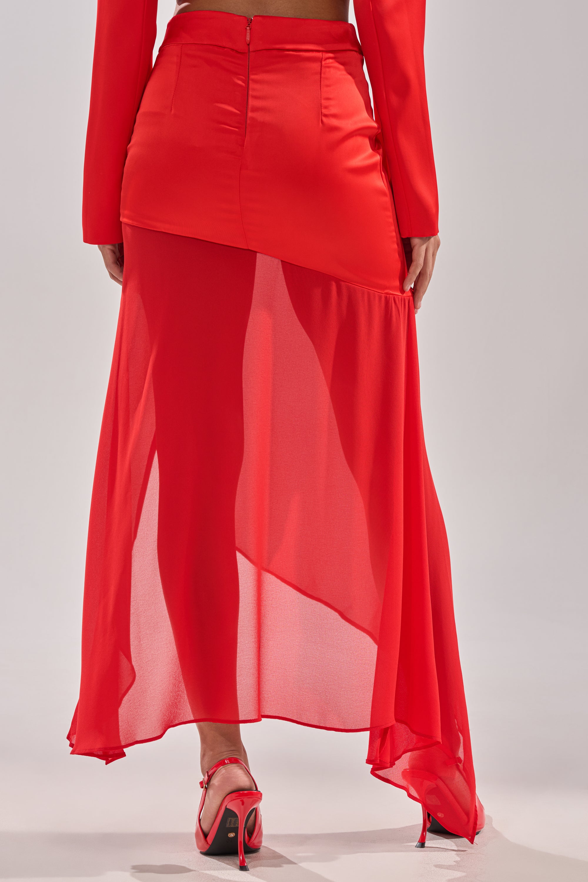Wearing the RED HOT ASYMMETRICAL MIDI SKIRT, featuring a high waist and sheer chiffon overlay, with red heeled sandals. The photo, shot from behind, emphasizes the skirt's bold silhouette. 4