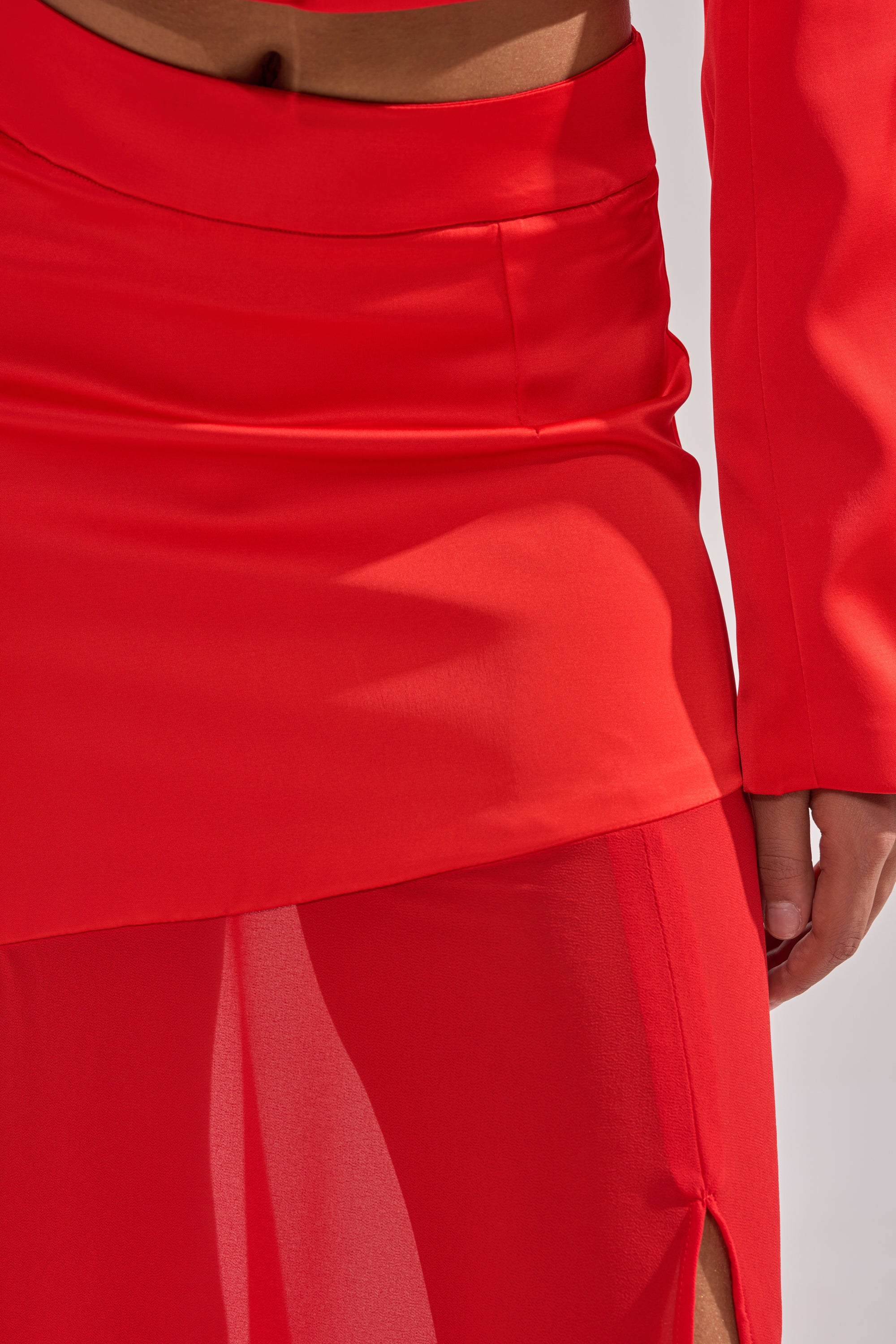 A close-up shows someone in a vibrant red outfit, featuring the RED HOT ASYMMETRICAL MIDI SKIRT with sheer fabric and a side slit, paired with a matching long-sleeve top. Their hand rests near their thigh. 5