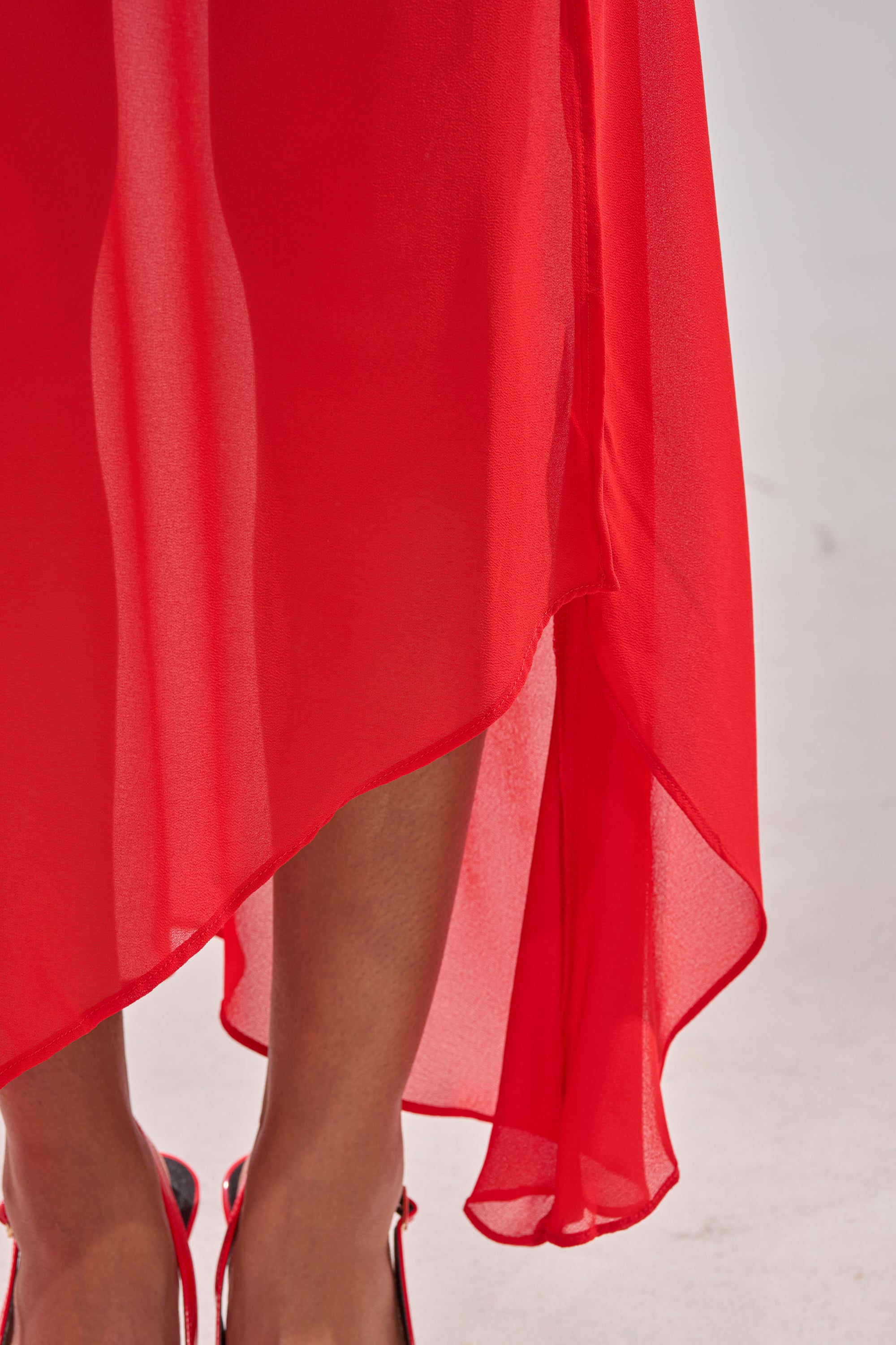 A close-up shows a person in the RED HOT ASYMMETRICAL MIDI SKIRT and red heels, the high-waisted, bright skirt’s asymmetrical hemline pops against a light gray background. 7