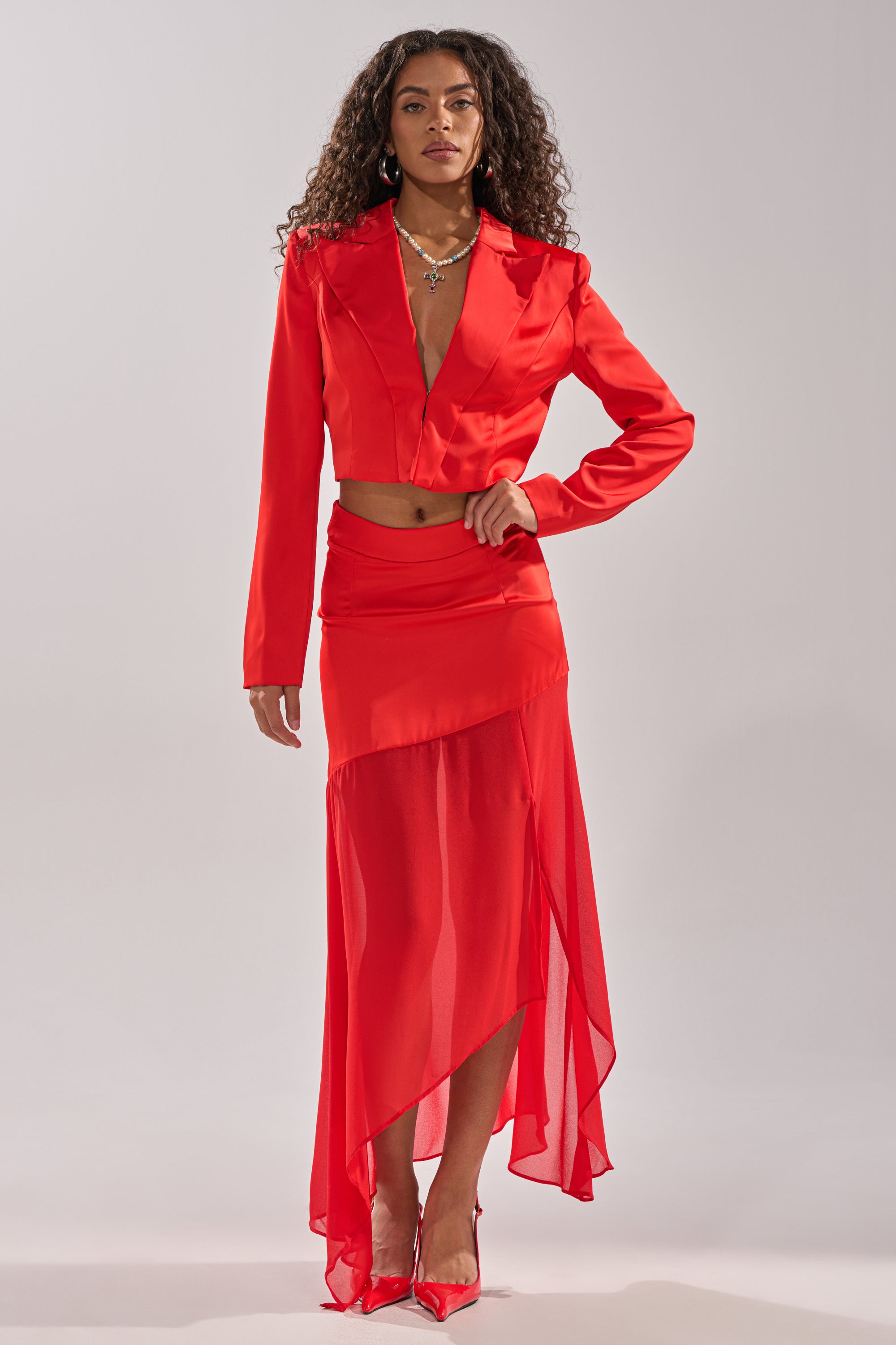 A woman faces forward in the RED HOT ASYMMETRICAL MIDI SKIRT set—a bold red, high-waist skirt with a sheer slit and cropped blazer. She has long curly hair, minimal jewelry, and wears red pointed heels. 8