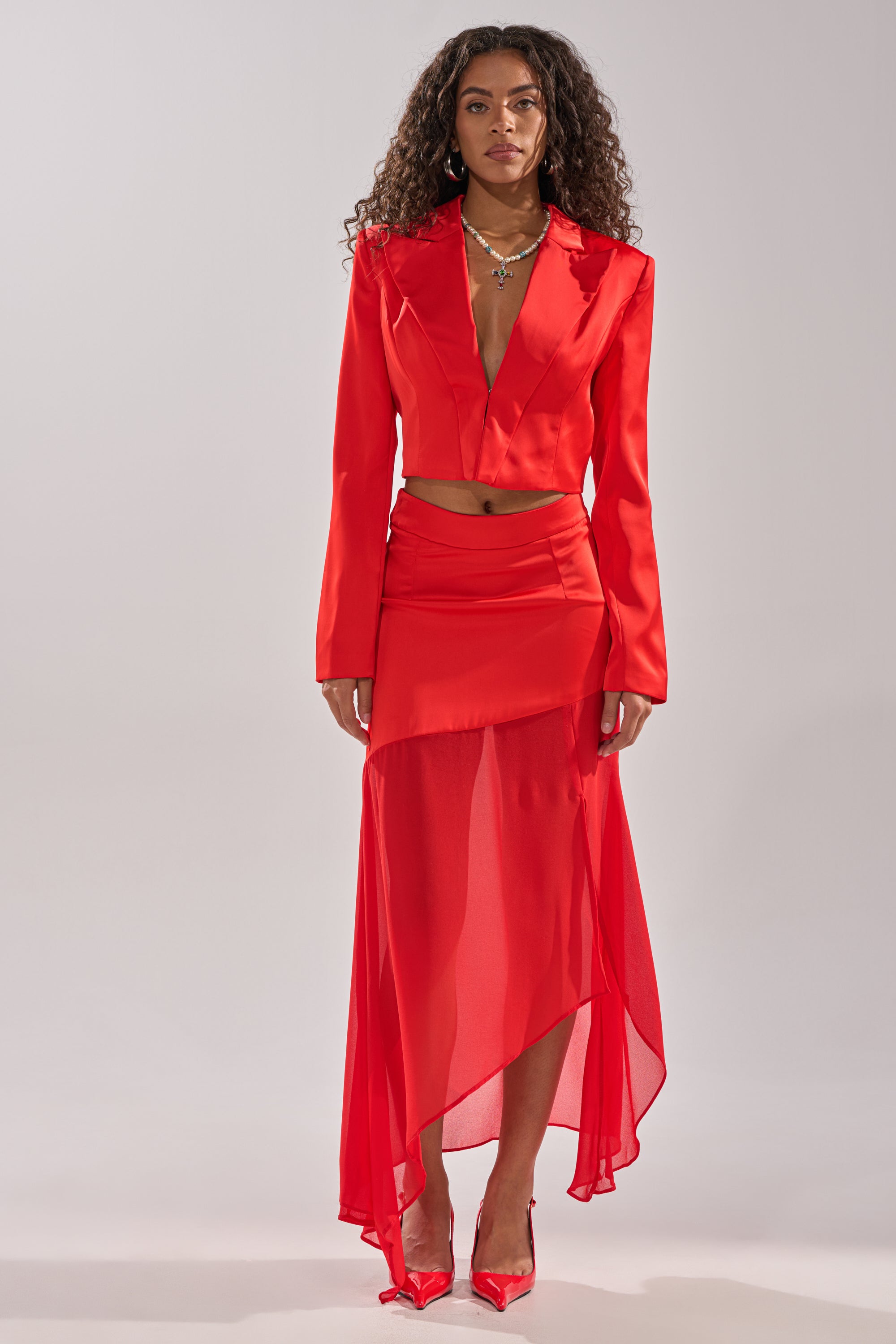 A woman faces forward in the RED HOT CROPPED SATIN BLAZER by AKIRA, styled with a matching sheer skirt, pointed red heels, curly hair, and layered necklaces for a bold, sophisticated look. 8