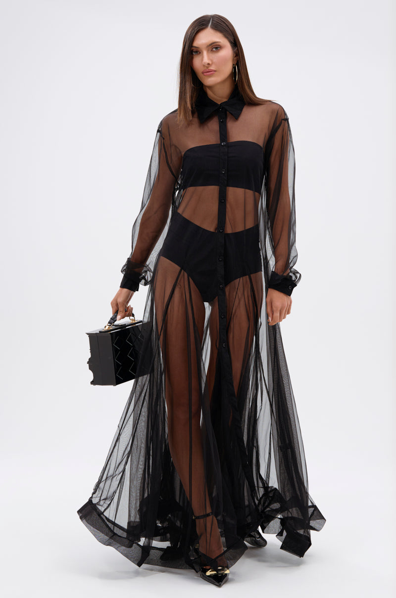 A woman in the COOL GIRL MESH LAYERING MAXI DRESS, featuring a sheer A-line silhouette, stands holding a black geometric handbag. She has straight brown hair and pointed black heels against a plain white background.