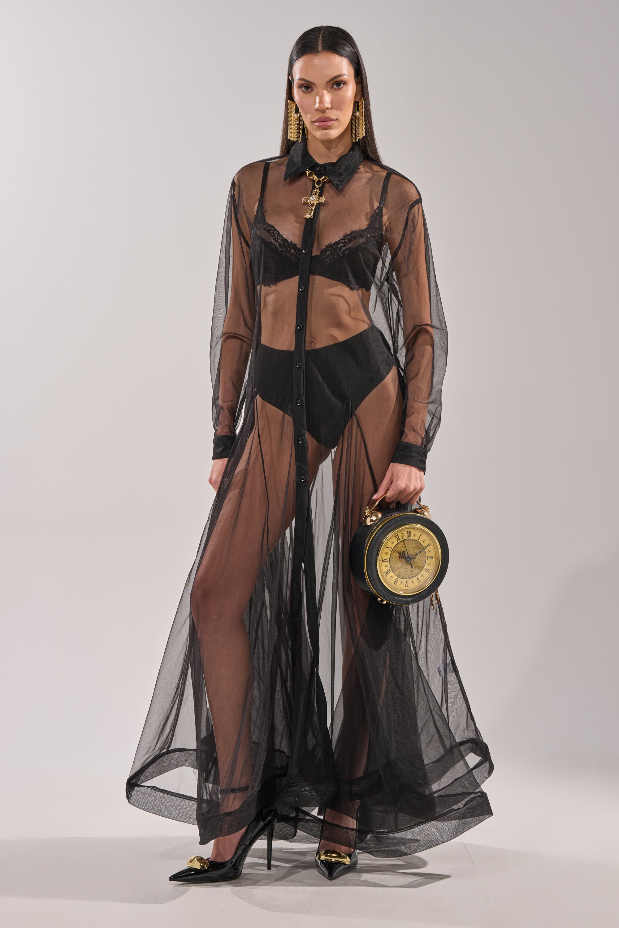 A woman models the COOL GIRL MESH LAYERING MAXI DRESS over black lingerie, styled with black heels, gold earrings, and a vintage clock-inspired round bag, posing against a plain light background.