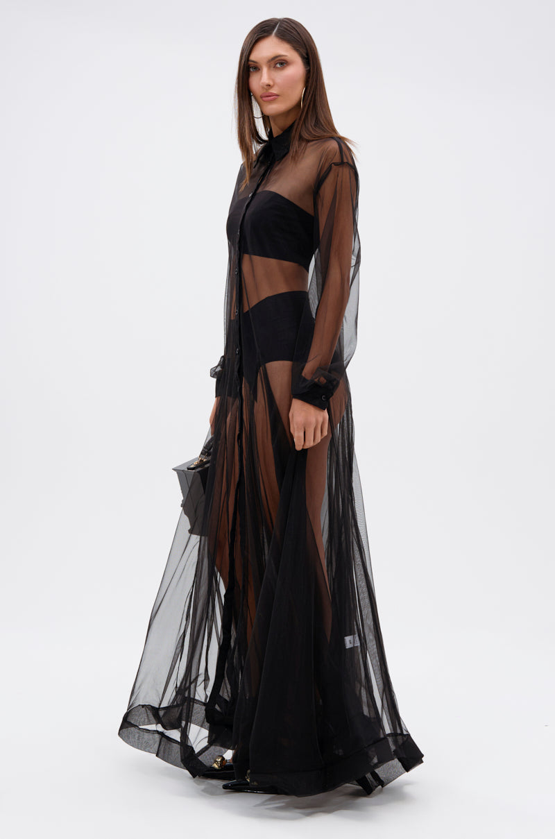 A woman with straight brown hair poses against a white background, wearing the COOL GIRL MESH LAYERING MAXI DRESS over black undergarments and holding a small black handbag. She looks slightly to the side.