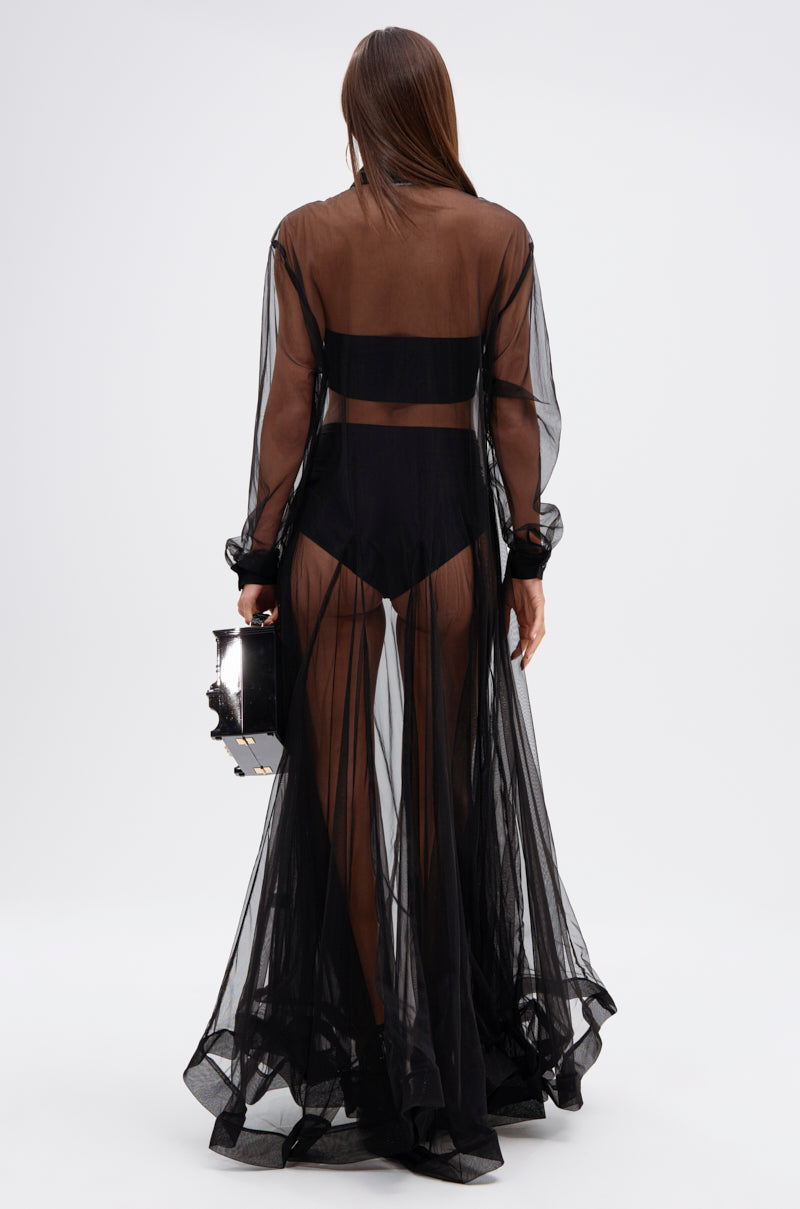 A person stands facing away in the COOL GIRL MESH LAYERING MAXI DRESS, showcasing its sheer mesh and A-line silhouette over black undergarments. Their straight brown hair cascades down as they hold a boxy black handbag against a plain light background.