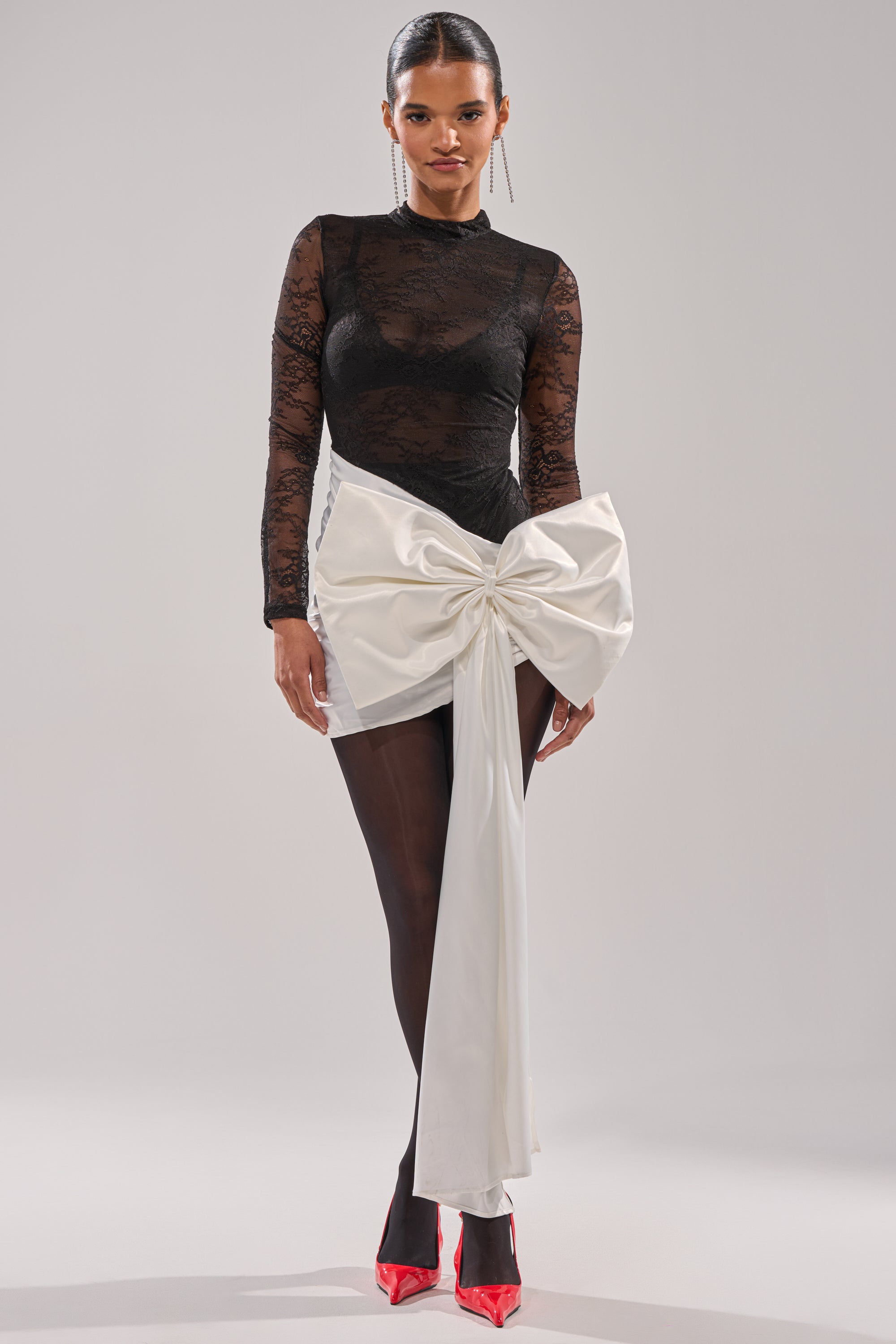 A woman wears the GIFT WRAPPED LONG SLEEVE LACE MINI DRESS with sheer tights, red pointed heels, and a white satin mini skirt featuring a sculptural front bow, standing against a plain light background.