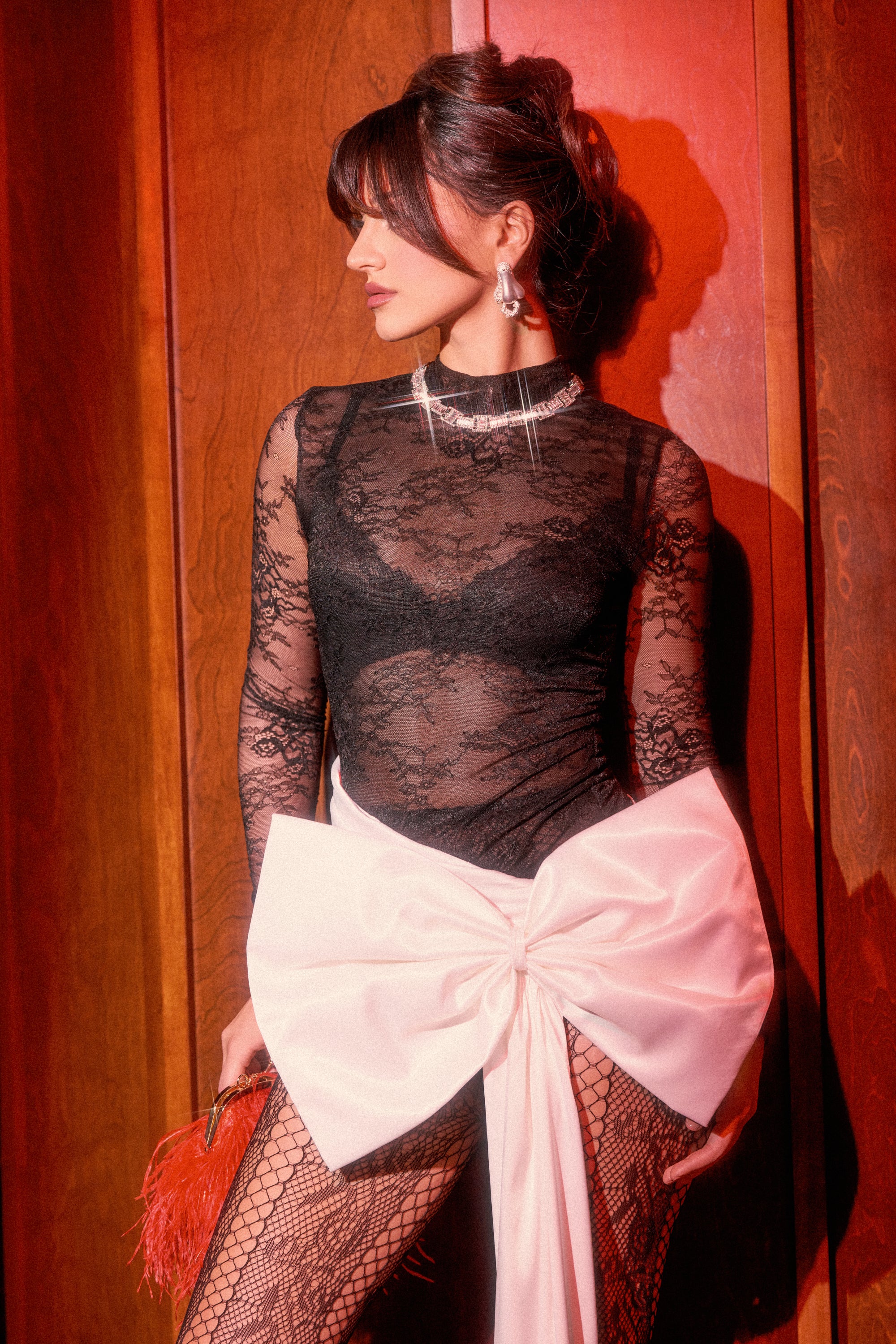 A woman stands against a wooden wall wearing the GIFT WRAPPED LONG SLEEVE LACE MINI DRESS styled like holiday party attire, featuring a large white bow at the waist. She accessorizes with pearl earrings and holds a red feathered clutch.