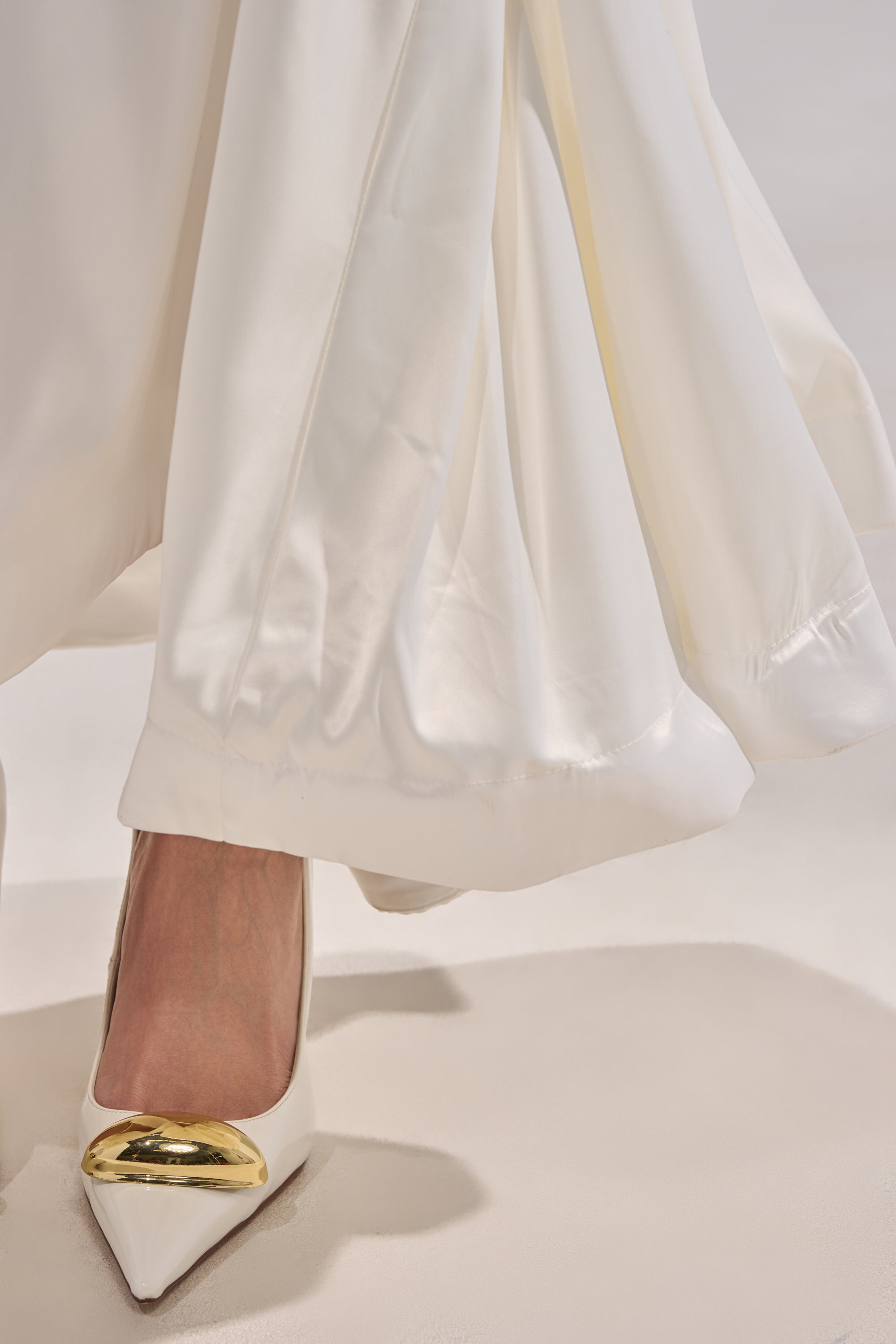 A close-up of a person wearing the SPIRITED AWAY MAXI SKIRT, crafted in white satin, paired with pointed white heels featuring shiny gold toe accents. The skirt's elegant fabric drapes gracefully toward the floor.