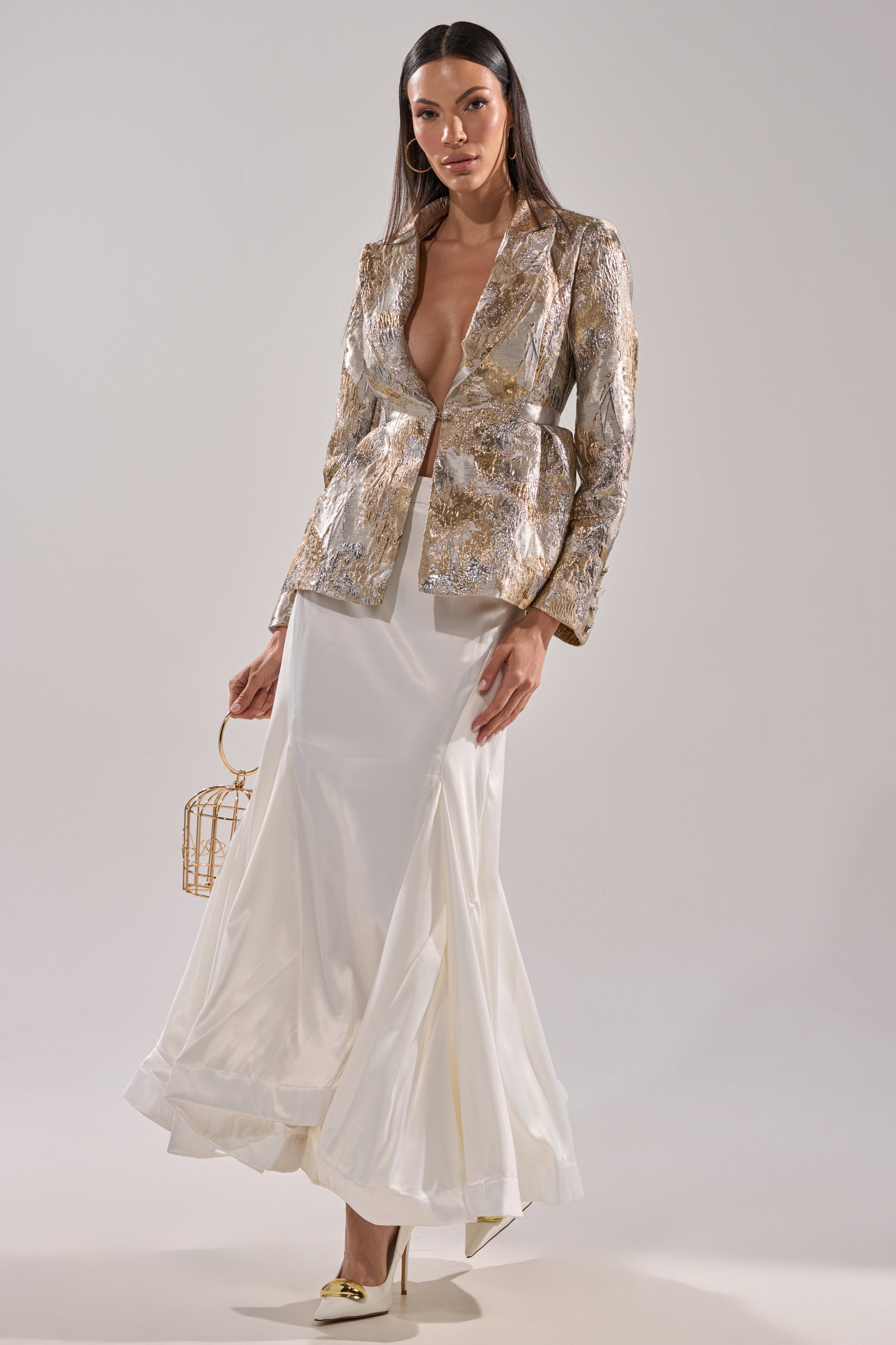 A woman stands confidently in the SPIRITED AWAY MAXI SKIRT, paired with a metallic gold patterned blazer, cream heels with gold accents, and a small gold geometric handbag. The background is plain and light-colored.
