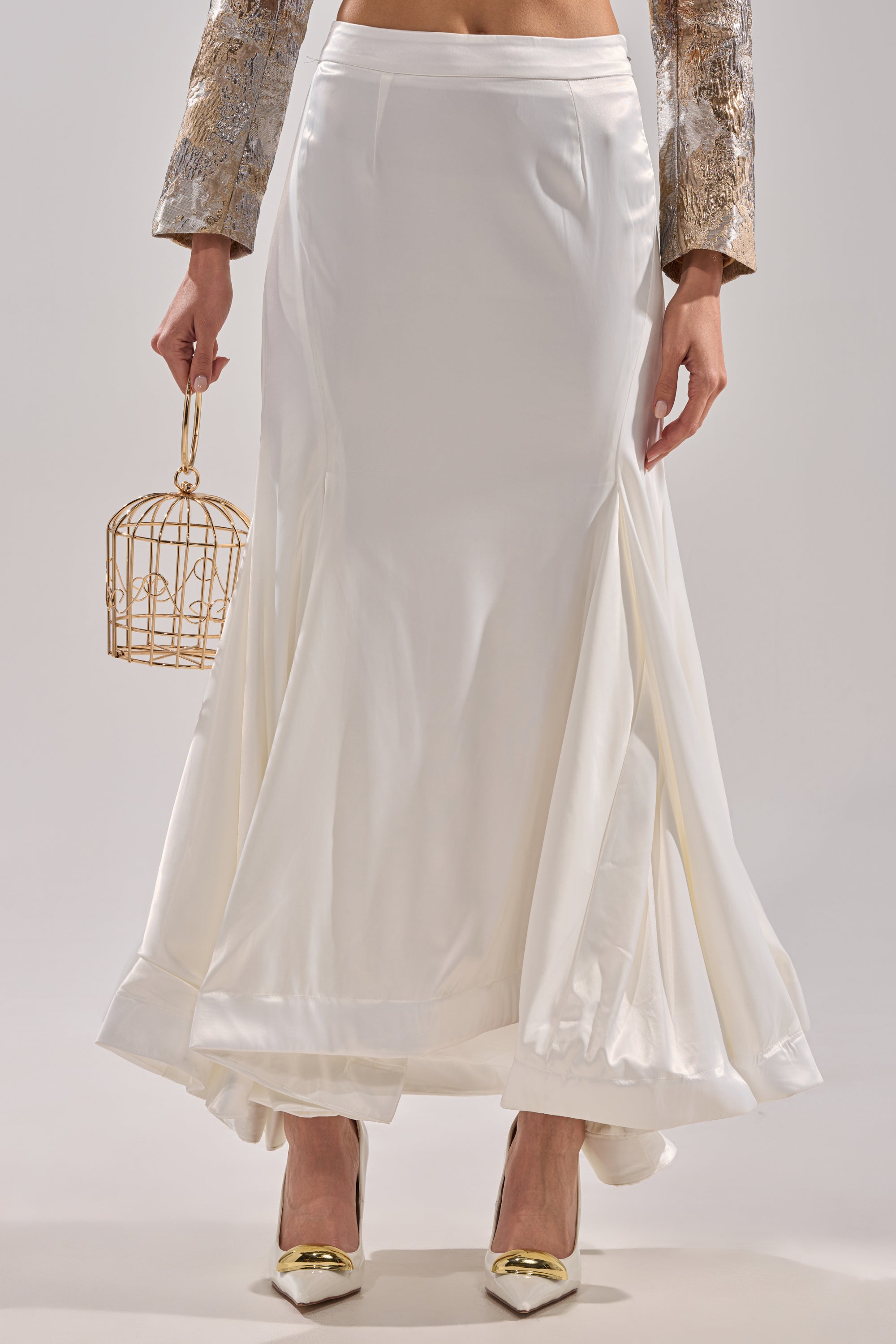 A woman wears the AKIRA Label SPIRITED AWAY MAXI SKIRT in white satin with flared panels, metallic gold heels, a gold-patterned jacket, and carries a small gold cage-shaped handbag.