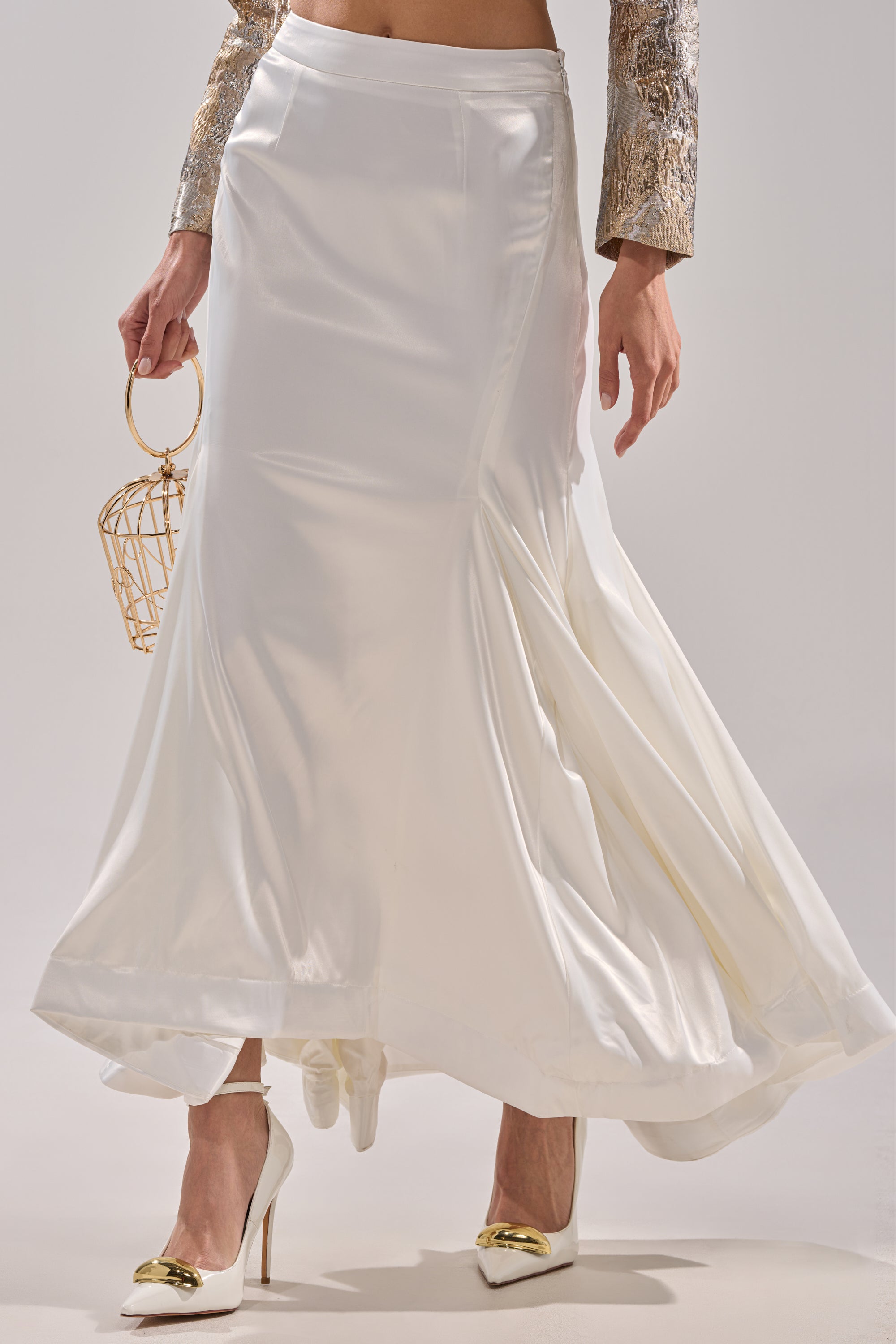 Wearing the SPIRITED AWAY MAXI SKIRT, white pointed-toe heels with gold tips, and a gold patterned top, the person holds a small gold wire handbag. The photo focuses on the chic lower half of this stylish look.