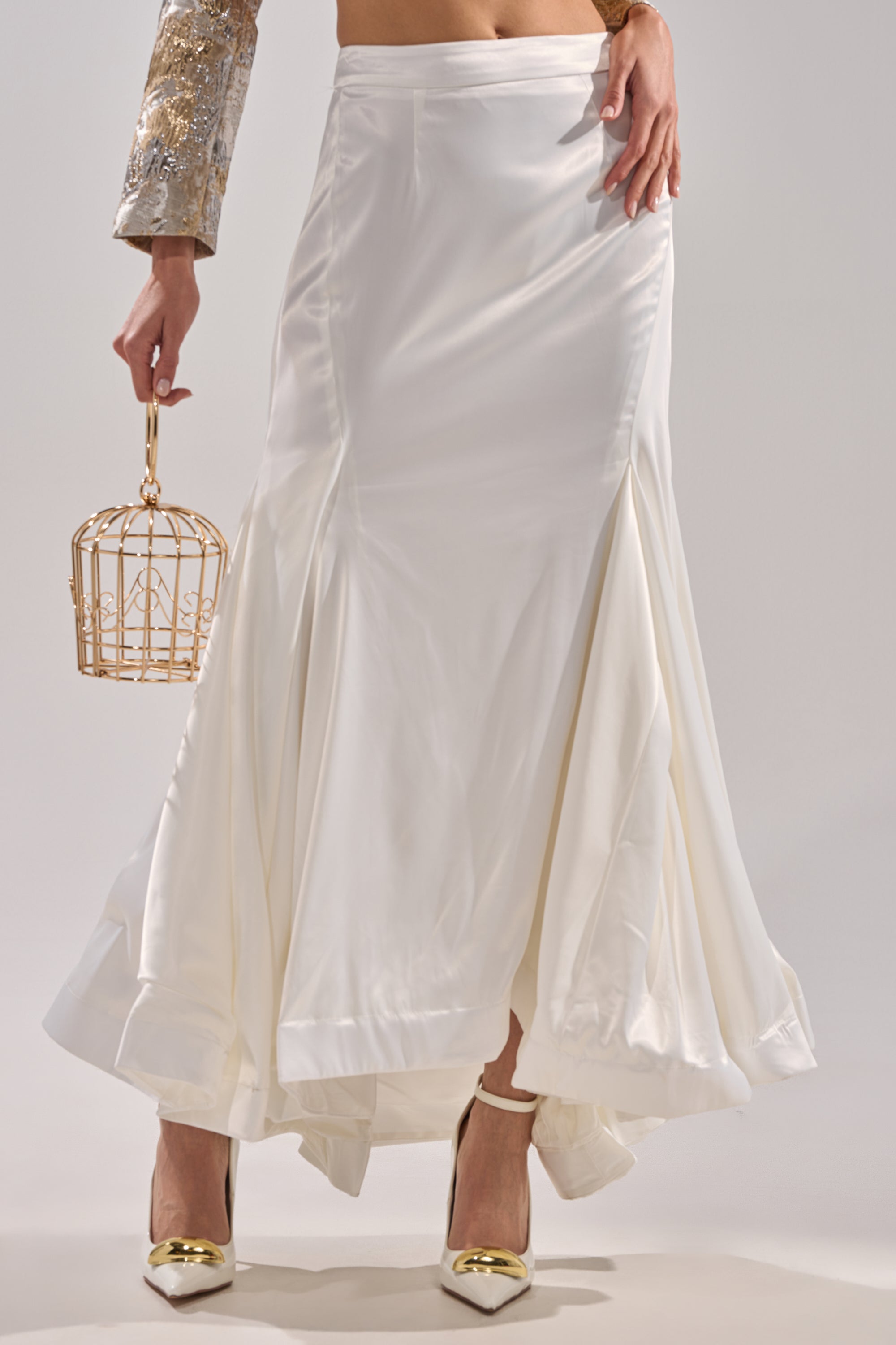 A person wearing the SPIRITED AWAY MAXI SKIRT and white heels holds a small gold wire cage. Visible are the skirt, legs, torso, and part of a gold-sequined sleeve.