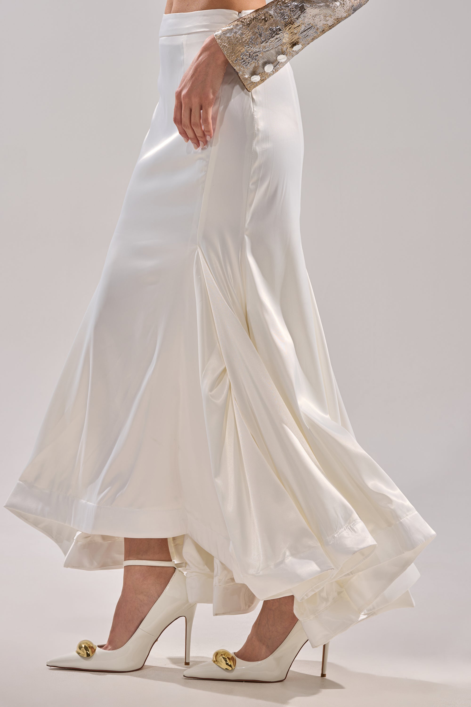 Wearing the SPIRITED AWAY MAXI SKIRT, a silver-sequined sleeve, and white high heels with gold accents, a person stands against a light background.