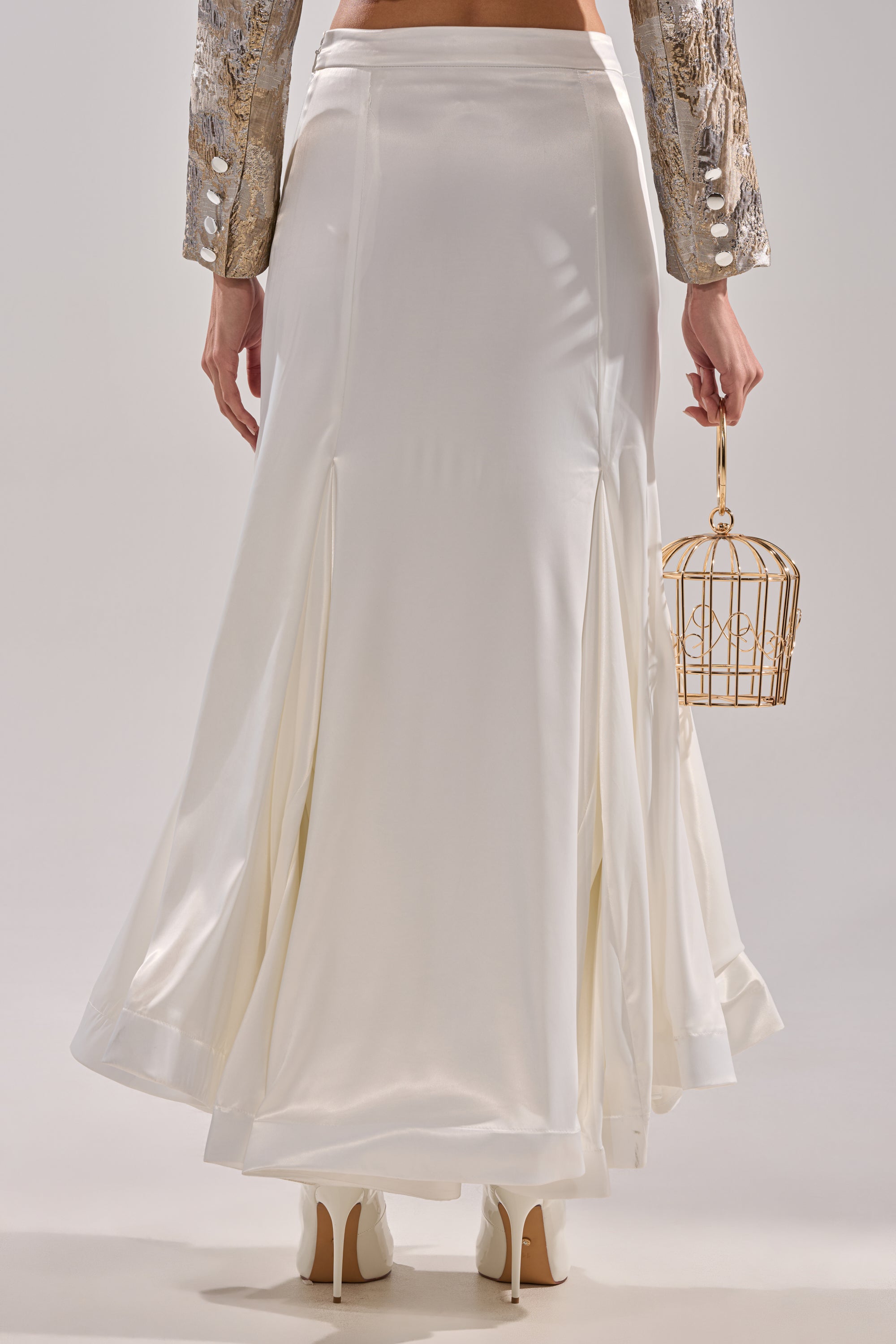 Wearing the SPIRITED AWAY MAXI SKIRT and white heels, a person holds a small gold birdcage-shaped handbag. The photo, taken from behind, captures only the lower half of their body.