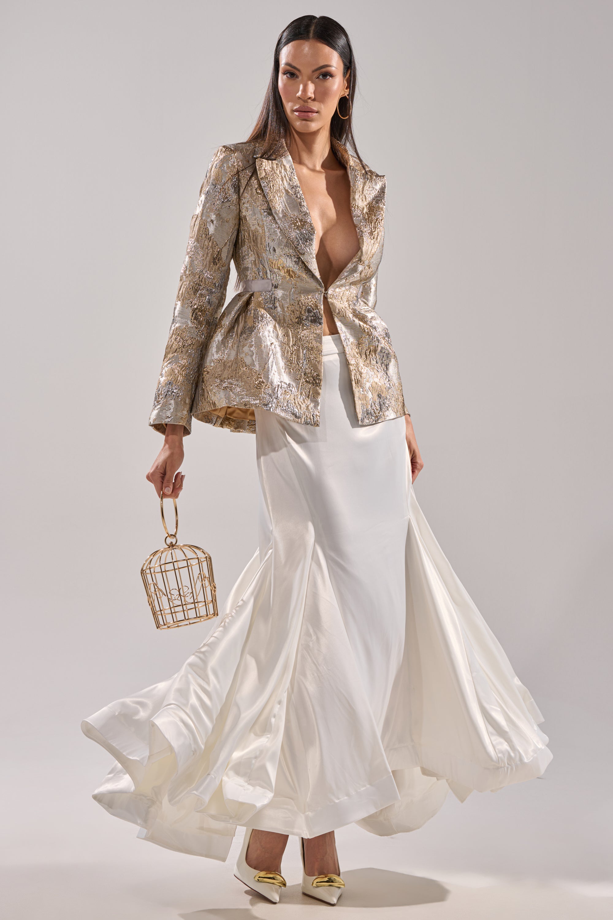A woman stands confidently in a metallic gold blazer with a deep neckline and the AKIRA Label SPIRITED AWAY MAXI SKIRT. She holds a small gold cage bag, wears pointed gold heels, and poses against a plain light-colored background.