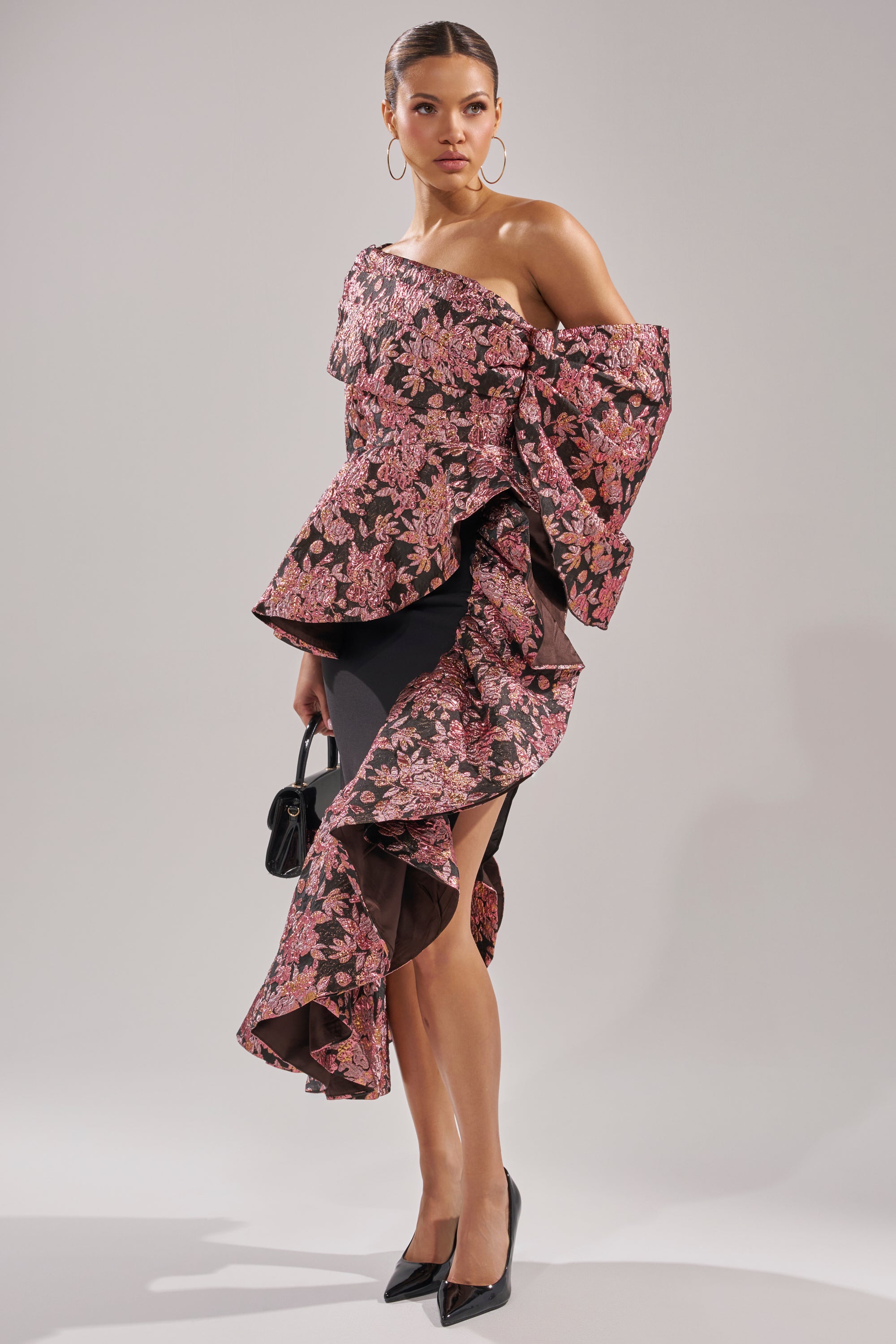 A woman poses against a plain background in the SO MUCH IN LOVE BROCADE MAXI SKIRT with a pink floral pattern, styled with black heels, a small black handbag, and large hoop earrings.