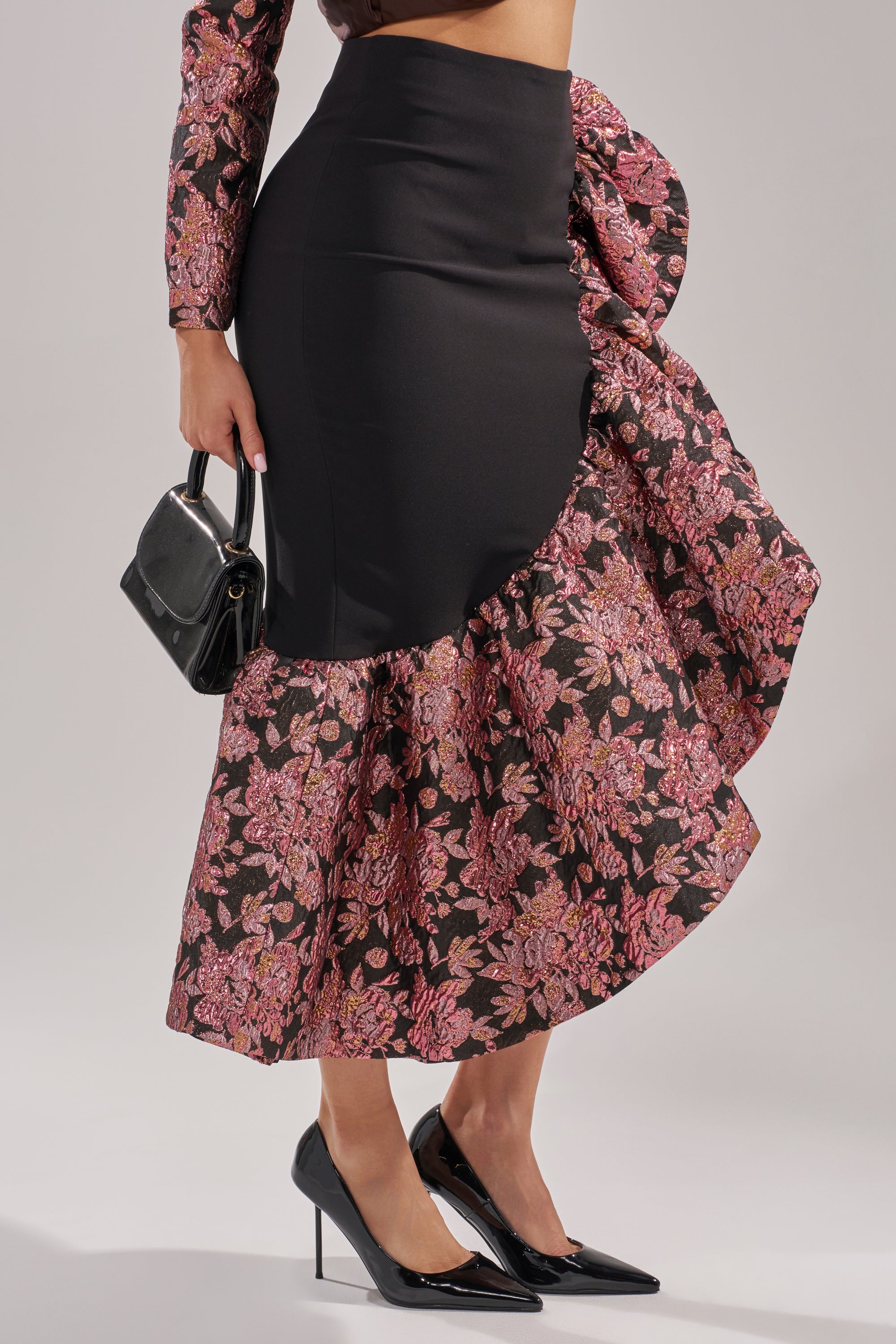 A woman models the SO MUCH IN LOVE BROCADE MAXI SKIRT, featuring a high-waisted fit and pink floral high-low ruffle hem, paired with a matching top, black patent heels, and a small black handbag—perfect for evening events.