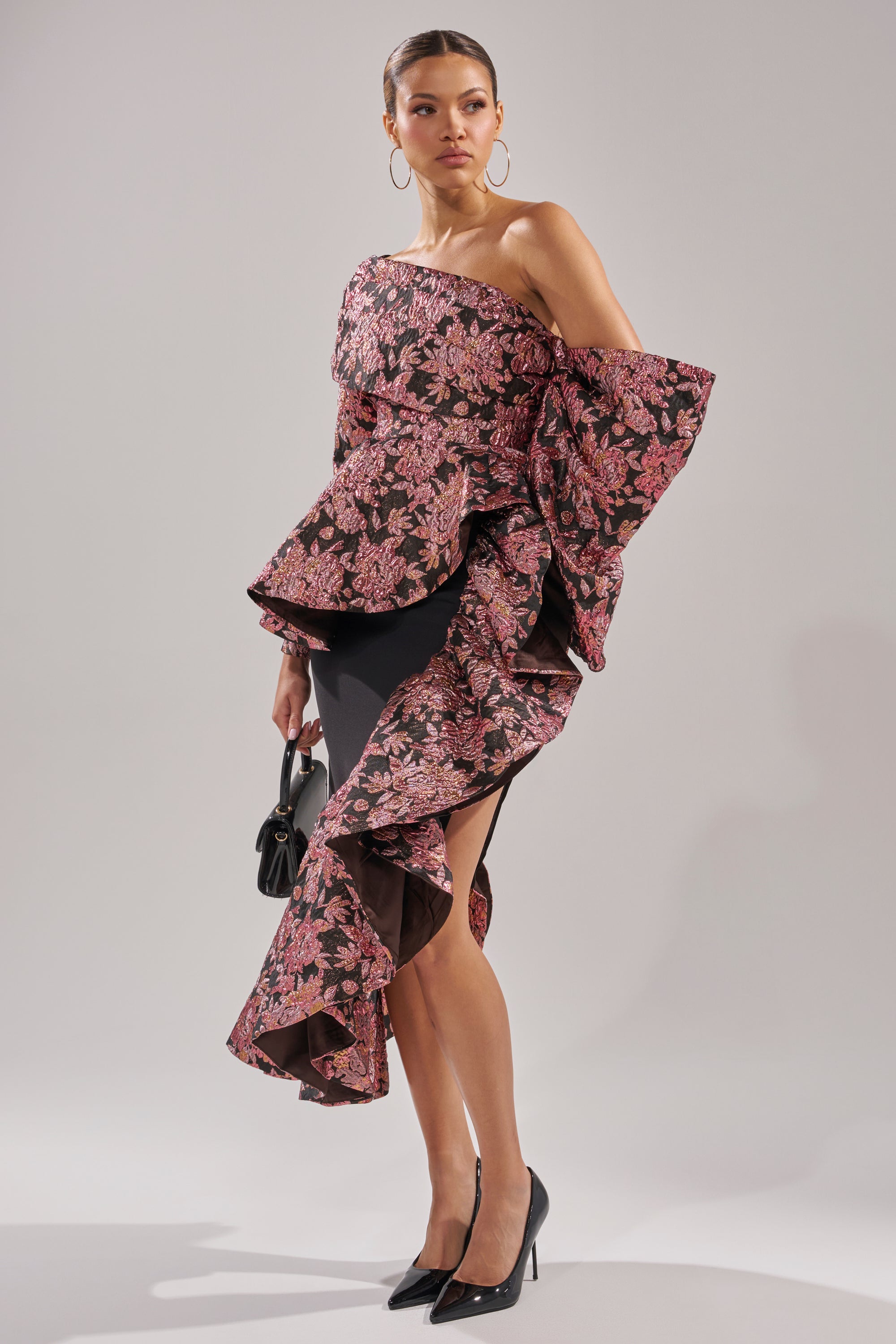A woman poses in the SO MUCH IN LOVE BROCADE MAXI SKIRT, featuring a pink and black floral pattern, dramatic high-low ruffle hemline, and high slit. She pairs it with black heels, gold hoop earrings, and a small black handbag.