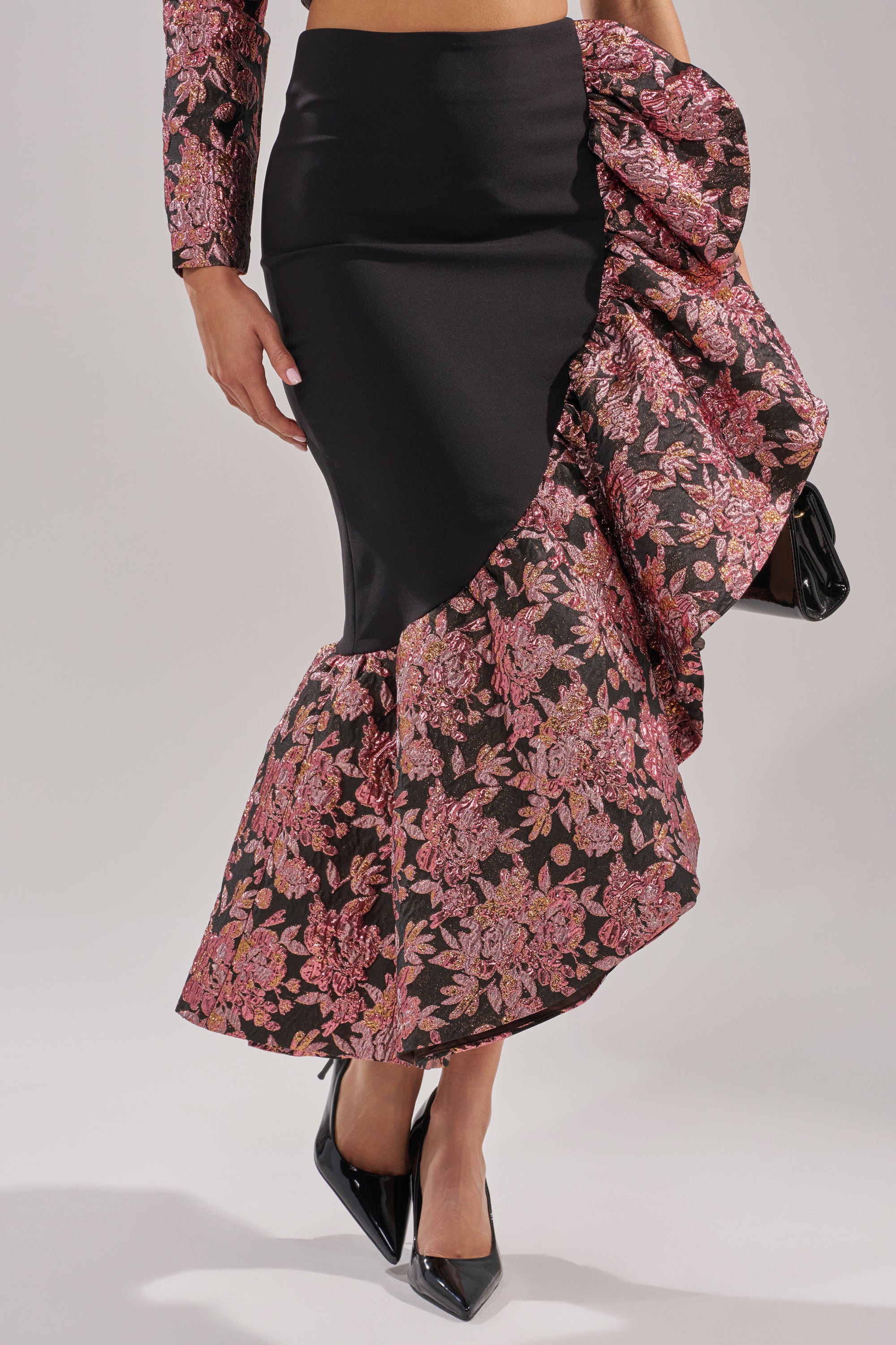 A woman wears the SO MUCH IN LOVE BROCADE MAXI SKIRT, a high-waisted black mermaid skirt with pink florals and a ruffle hem, paired with a matching top and black heels—ideal for a chic holiday party look.