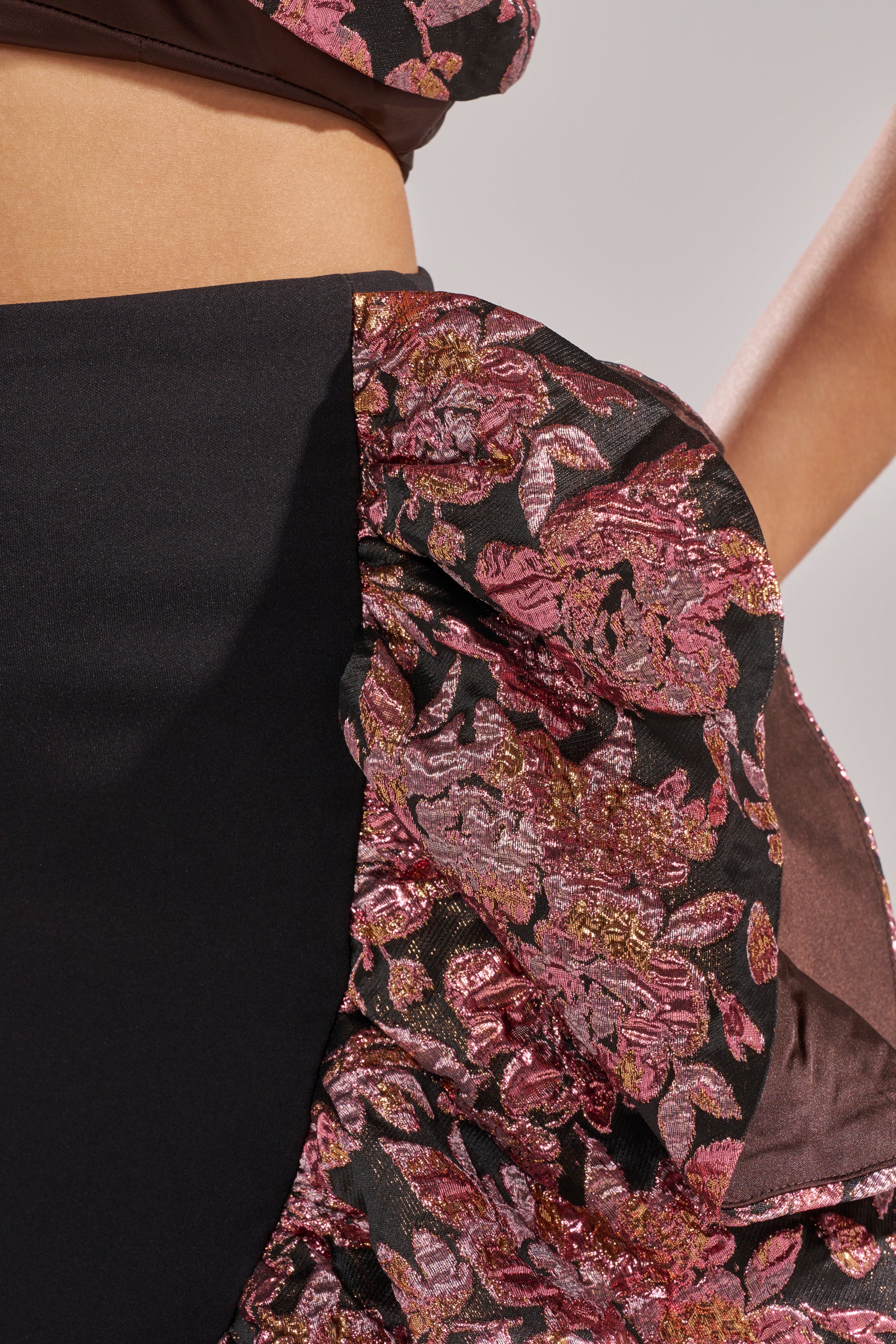 Close-up of a person wearing the SO MUCH IN LOVE BROCADE MAXI SKIRT, featuring textured, shiny pink and gold floral ruffles and a high-low hem. The image highlights the skirt’s festive fabric details, plus the person's bare midriff and arm.