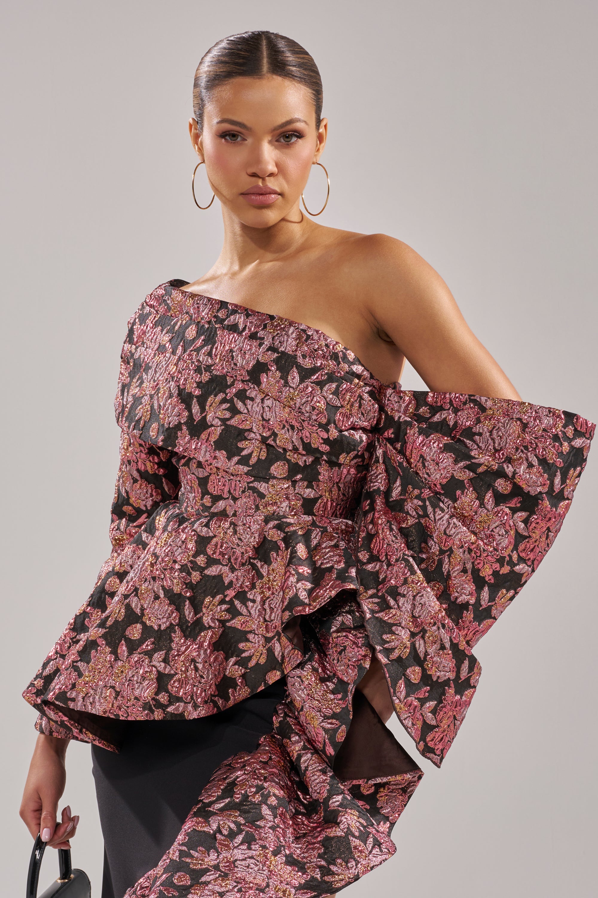 A woman wears the SO MUCH IN LOVE BROCADE BLOUSE, featuring bold metallic pink floral patterns on black, paired with hoop earrings and her hair styled back.