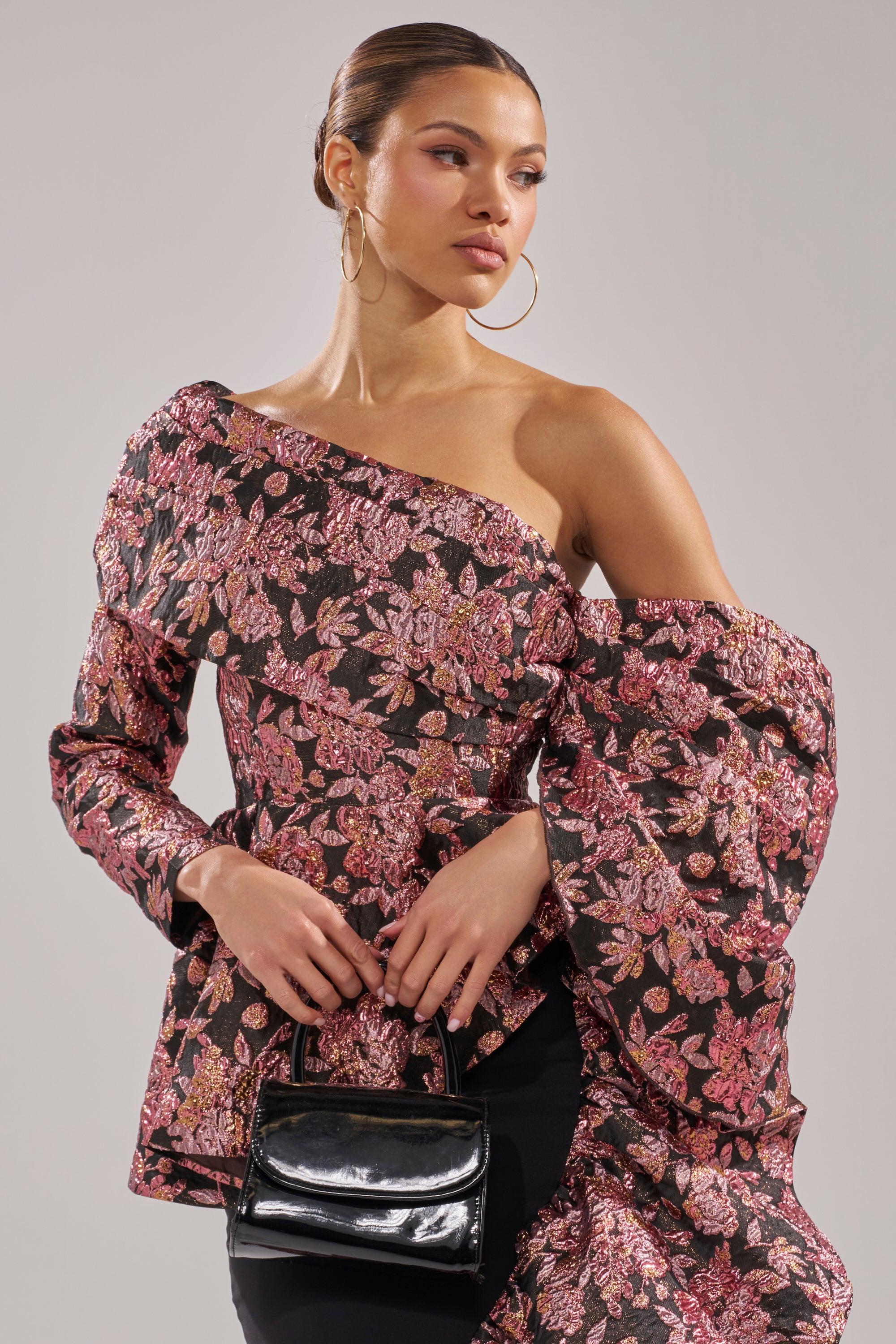 A woman wears the SO MUCH IN LOVE BROCADE BLOUSE, featuring a metallic floral design and voluminous sleeves. She accessorizes with large hoop earrings, a small black handbag, and has her hair pulled back while looking to the side.