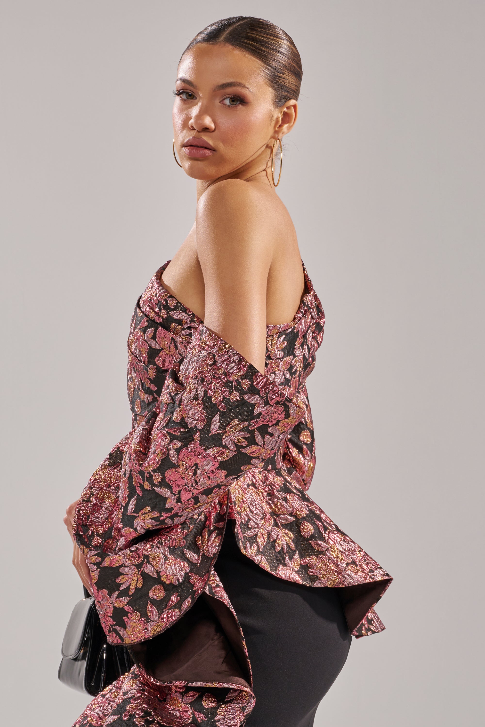 A woman wearing hoop earrings and slicked-back hair poses in the SO MUCH IN LOVE BROCADE BLOUSE, featuring pink and gold patterns, while holding a black clutch against a neutral background.