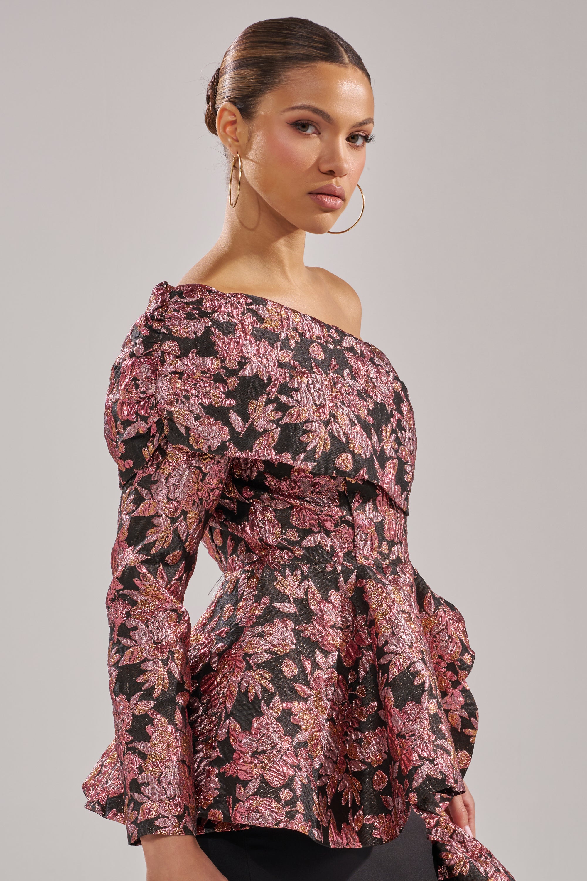 A woman with slicked-back hair and large hoop earrings wears the SO MUCH IN LOVE BROCADE BLOUSE—an off-the-shoulder, long-sleeve top with a pink and gold floral pattern—standing against a plain light gray background.