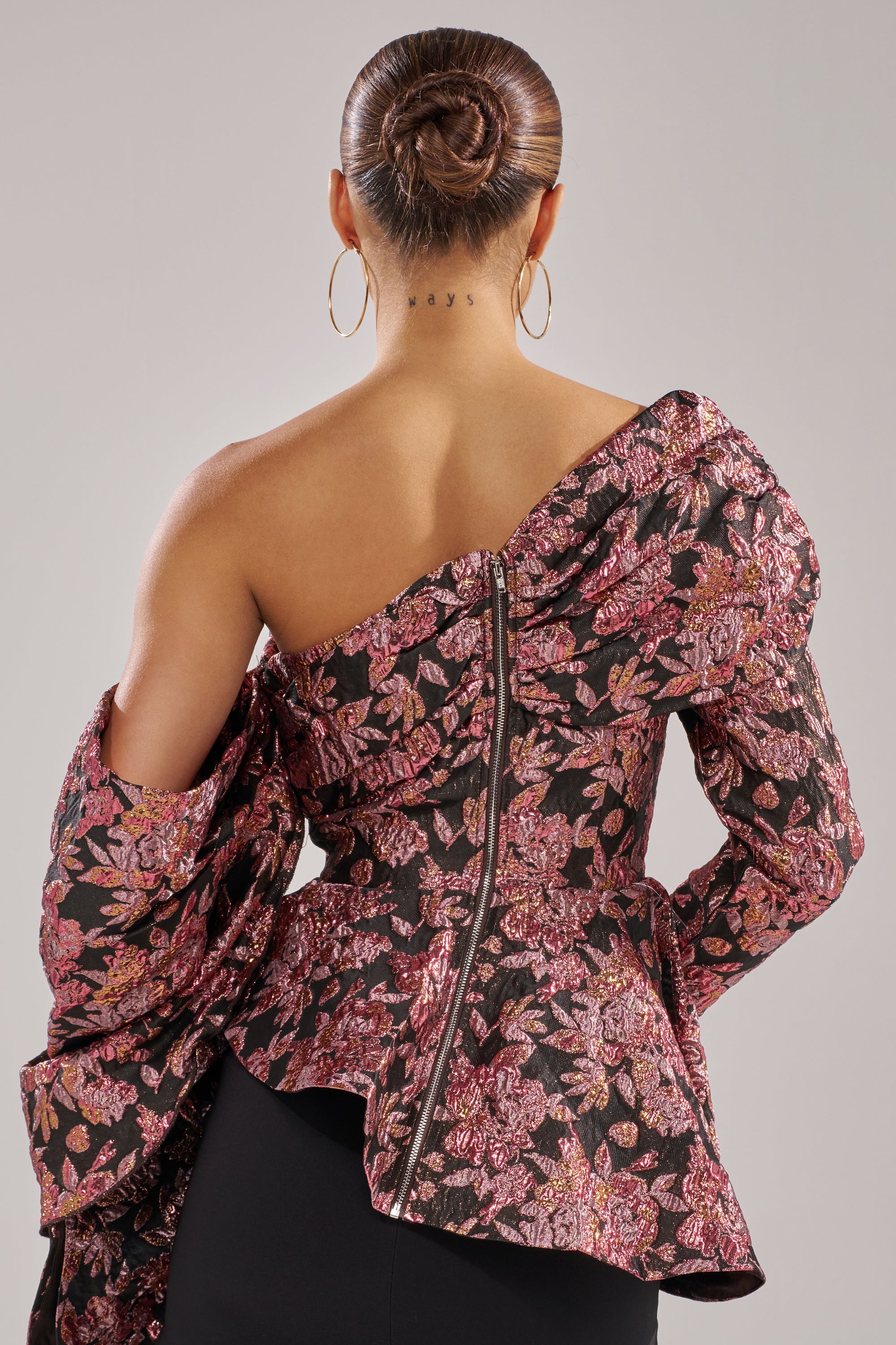 A person with hair in a neat bun, sporting large hoop earrings and the SO MUCH IN LOVE BROCADE BLOUSE with a back zipper, stands facing away. A "STAY" tattoo is visible on the back of their neck.
