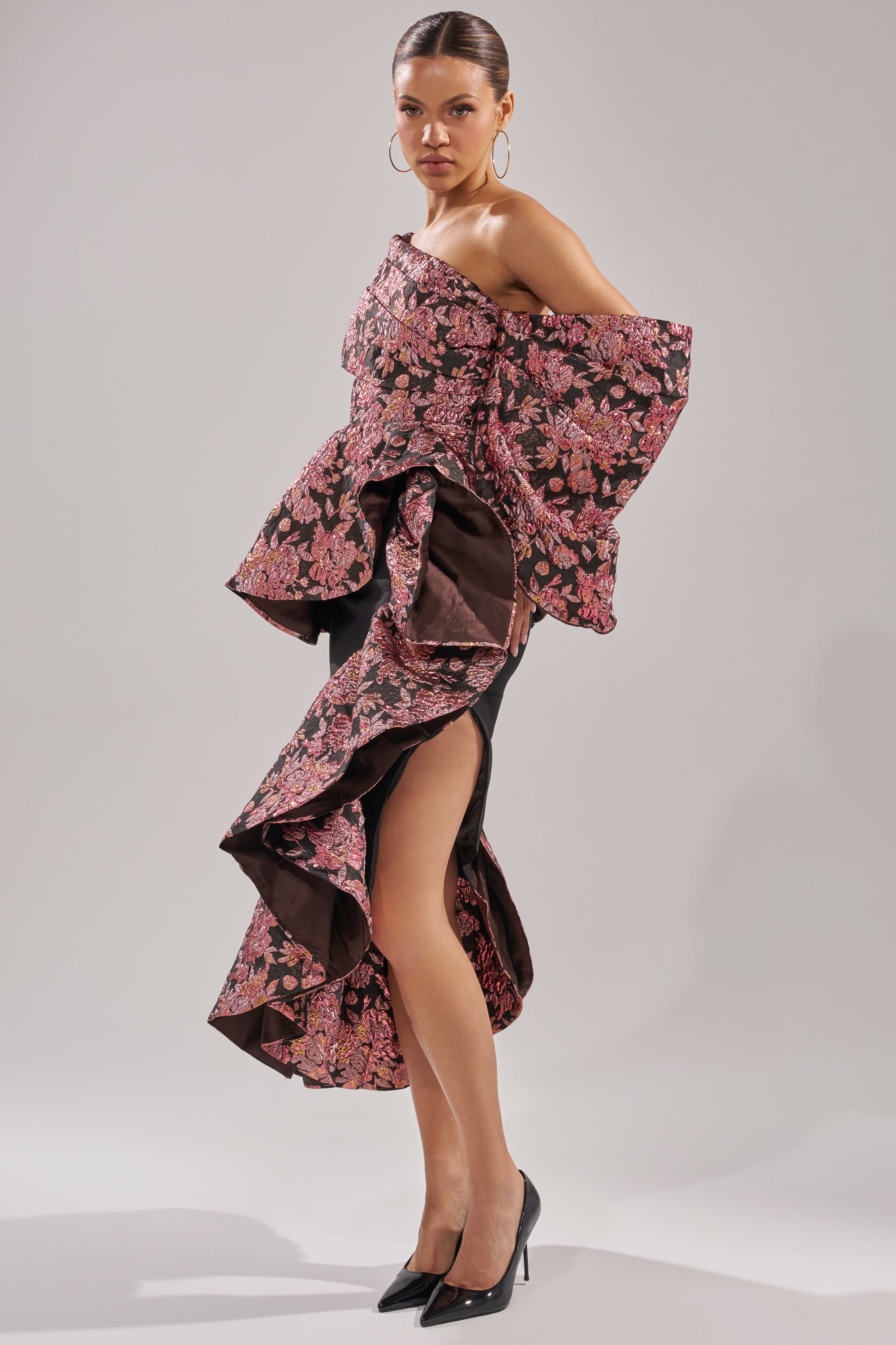 A woman poses in the SO MUCH IN LOVE BROCADE BLOUSE, featuring dramatic off-the-shoulder ruffles and a pink and brown floral pattern, styled with black high heels and hoop earrings against a plain light background.