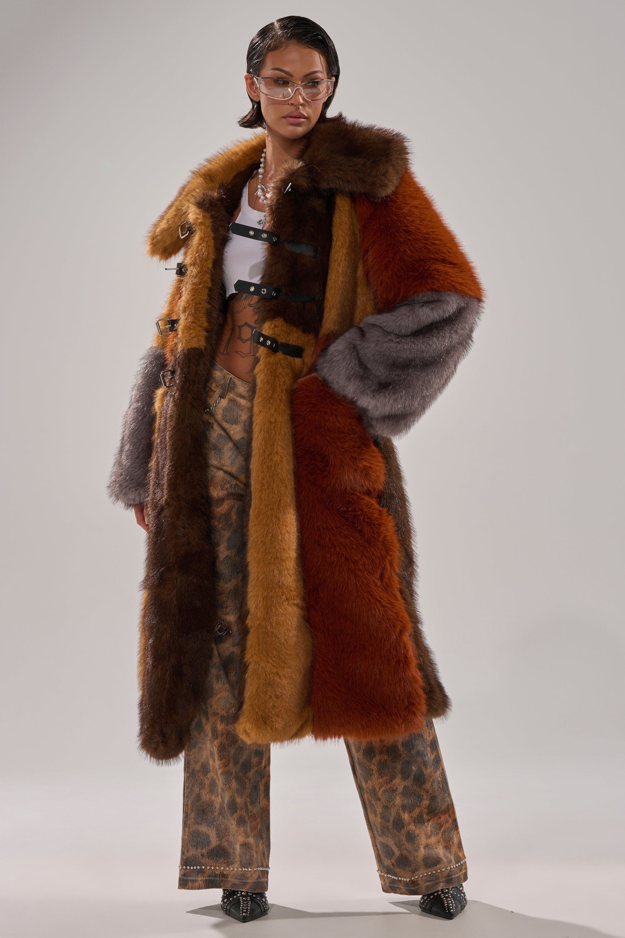 A person wears the WILD CARD PATCHWORK FAUX FUR coat, featuring bold brown, orange, and gray panels over a white crop top and animal print pants, styled with glasses and hoop earrings.