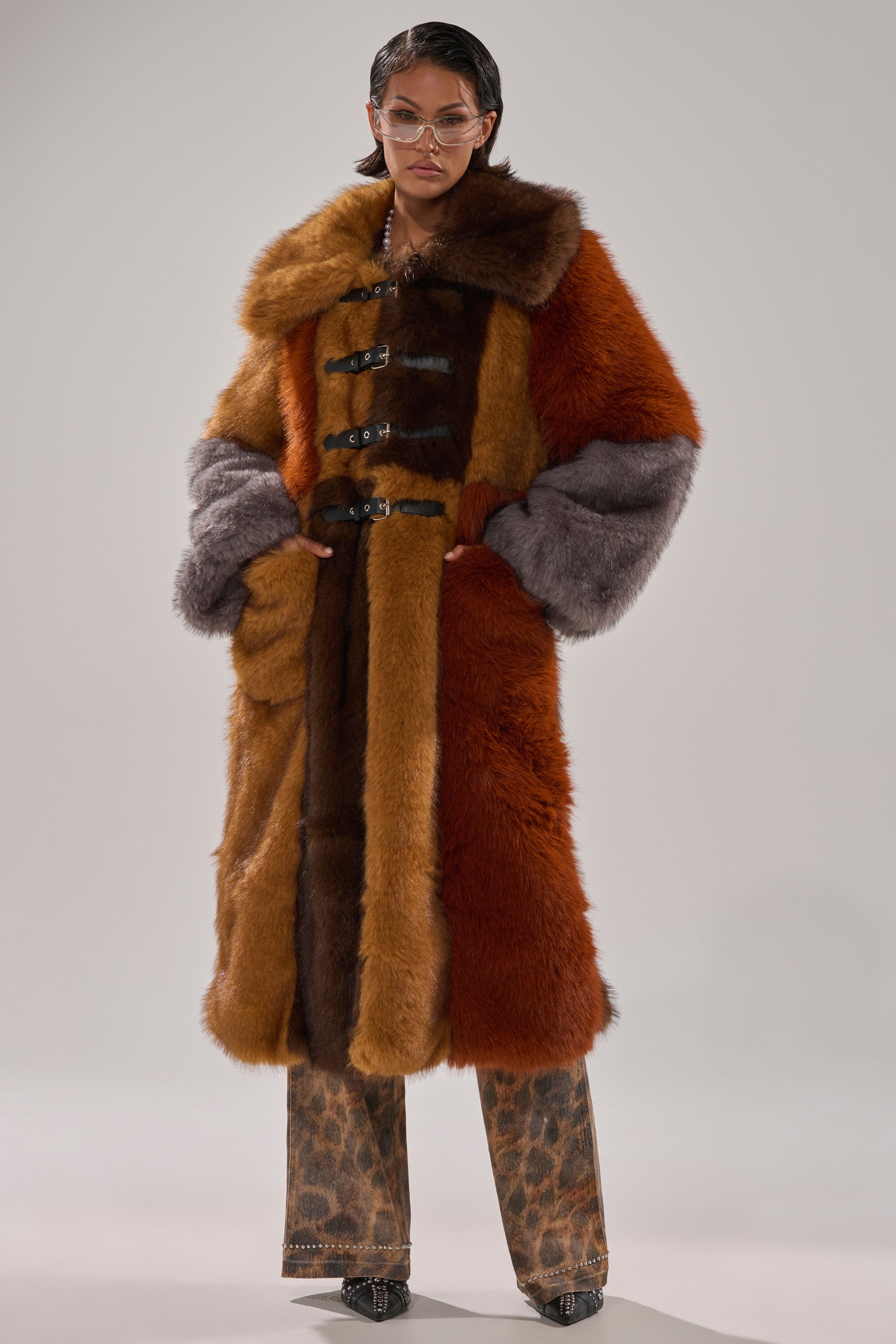 A person wears the WILD CARD PATCHWORK FAUX FUR coat in multicolored brown, orange, and gray, paired with animal print pants, sunglasses, and black shoes, standing against a plain background with hands in their coat pockets.